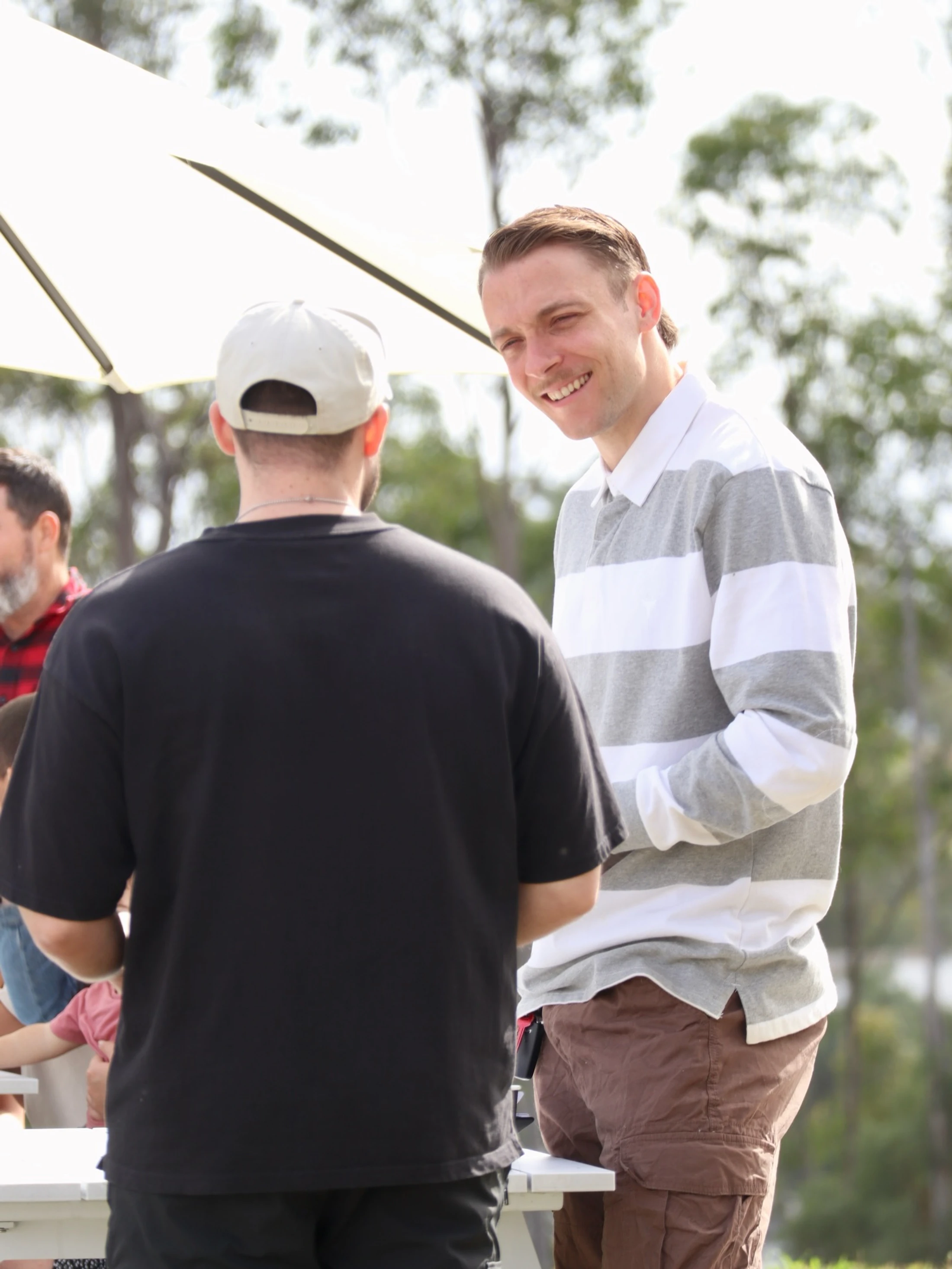 A man in a striped polo shirt smiles as he talks to another man in a black shirt and a white cap, who has his back to the camera. They are standing outdoors.