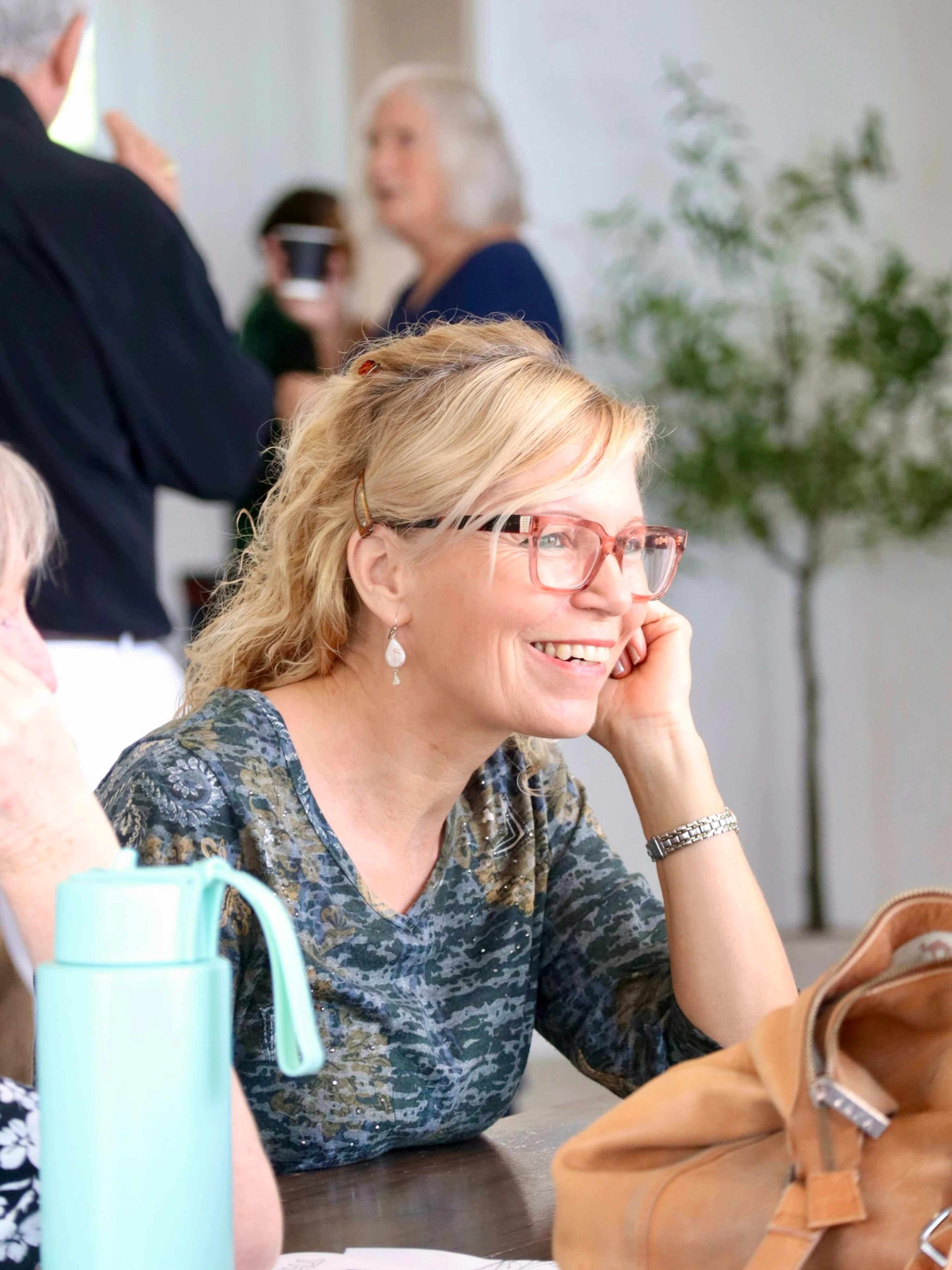 A smiling blonde woman with pink-rimmed glasses sits at a table, resting her chin on her hand as she listens during a social event.