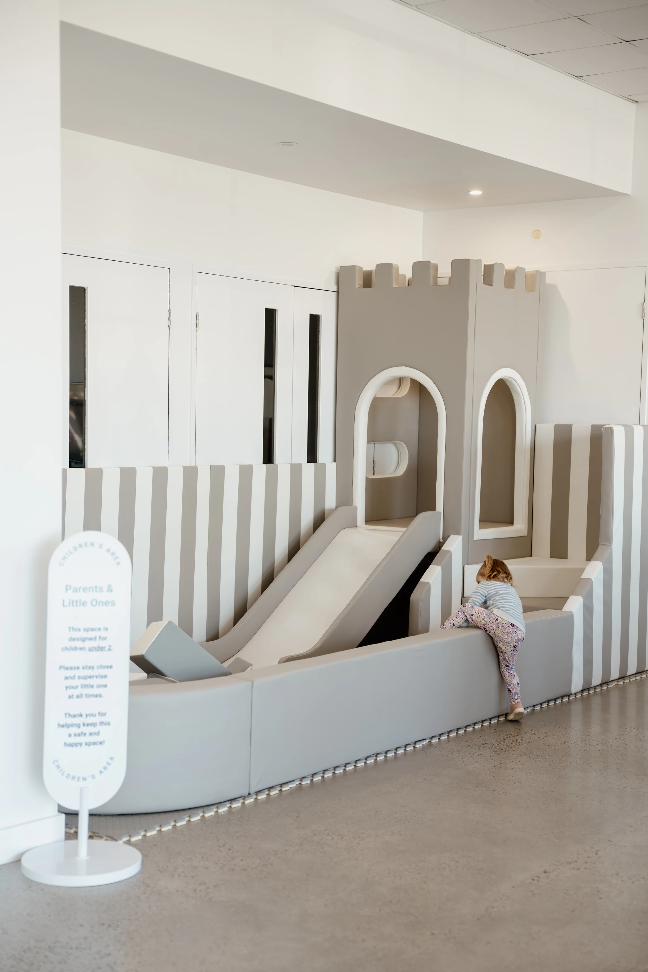 A young girl climbs into a padded, neutral-colored soft play area featuring a small castle with a slide. A sign nearby labels it a Children's Area.