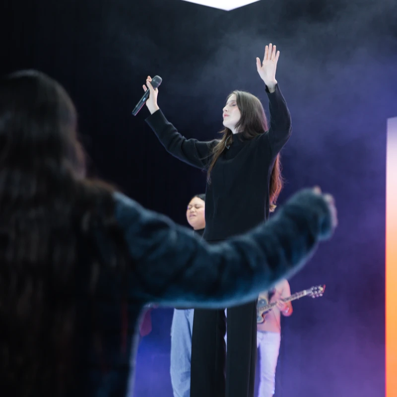 A woman in a black outfit stands on a brightly lit stage, singing into a microphone with her hand raised. A person in the foreground also has their arms up.