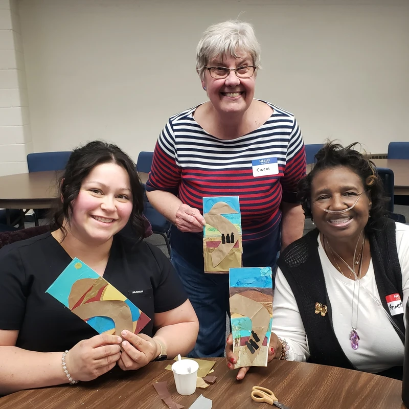 Three smiling women of different ages and races pose at a table in a classroom, proudly showing off the colorful paper collages they created.