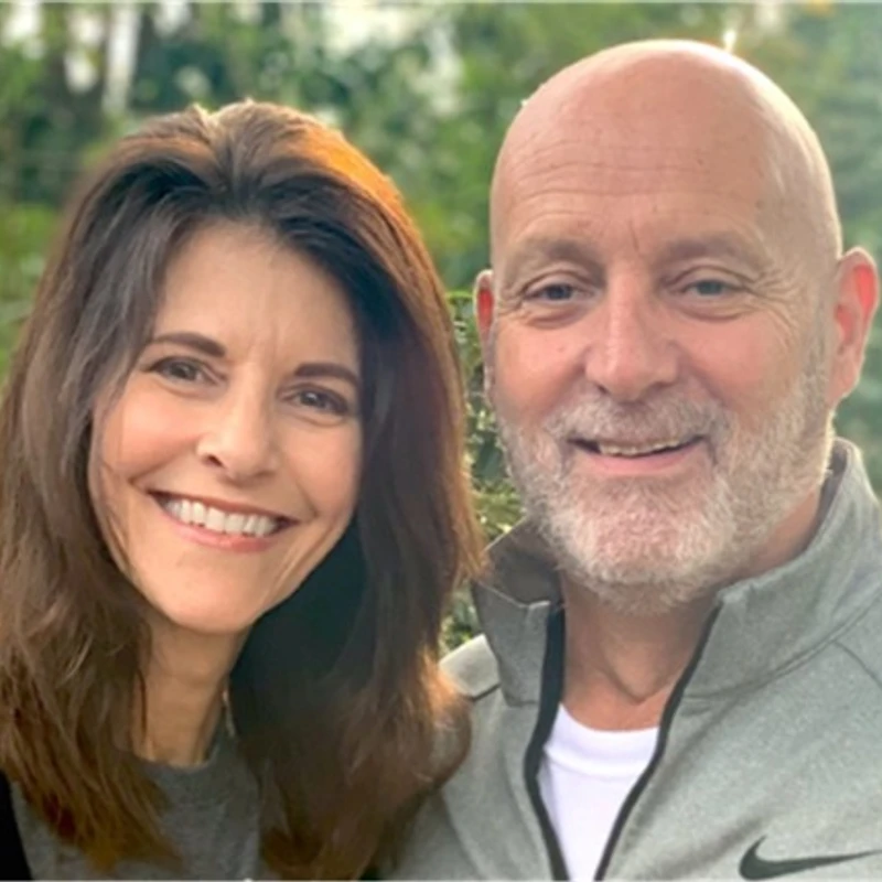 A smiling middle-aged couple stands close together outdoors. The woman has shoulder-length brown hair, and the man is bald with a gray beard.