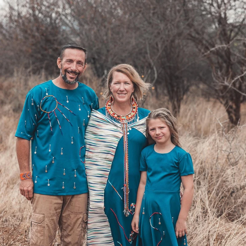 A family of three, a man, woman, and young girl, stand in a grassy field wearing matching teal outfits with traditional beaded decorations.