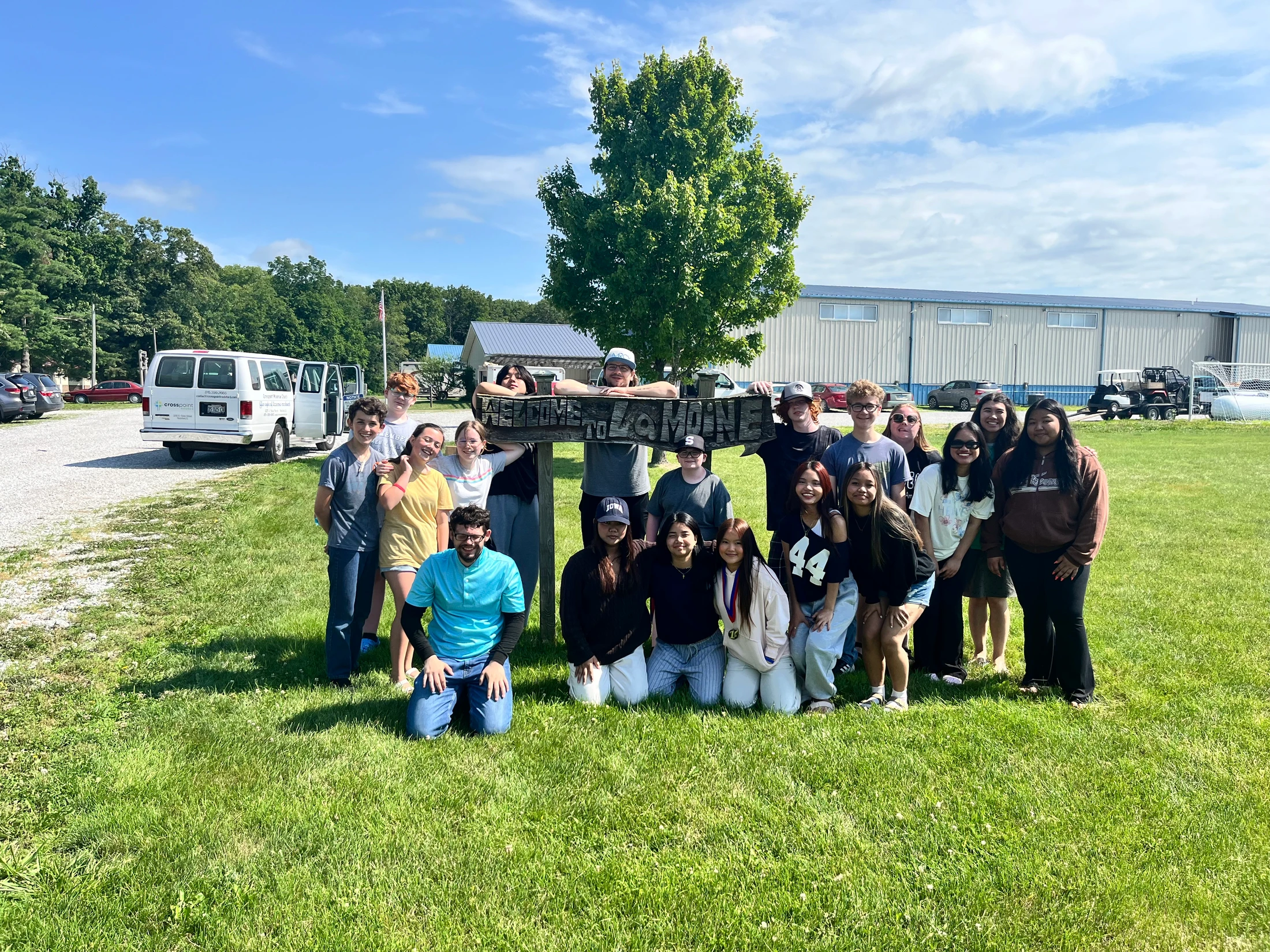 A diverse group of young people smiles for a photo, gathered around a rustic wooden welcome sign on a bright, sunny day in a grassy field.