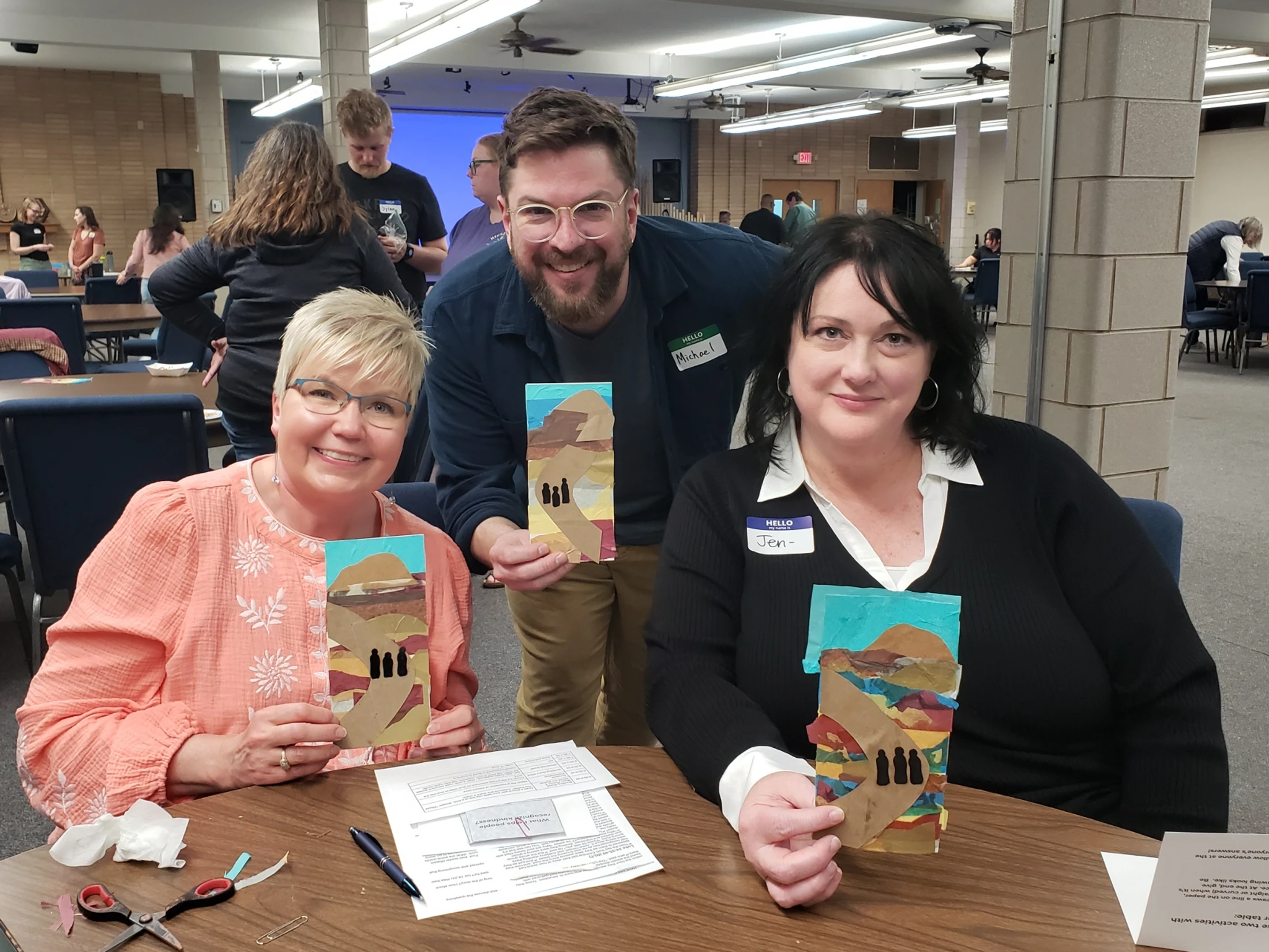 Three smiling adults, two women sitting and one man standing, pose for a photo at a table, holding up small paper collage artworks they created.