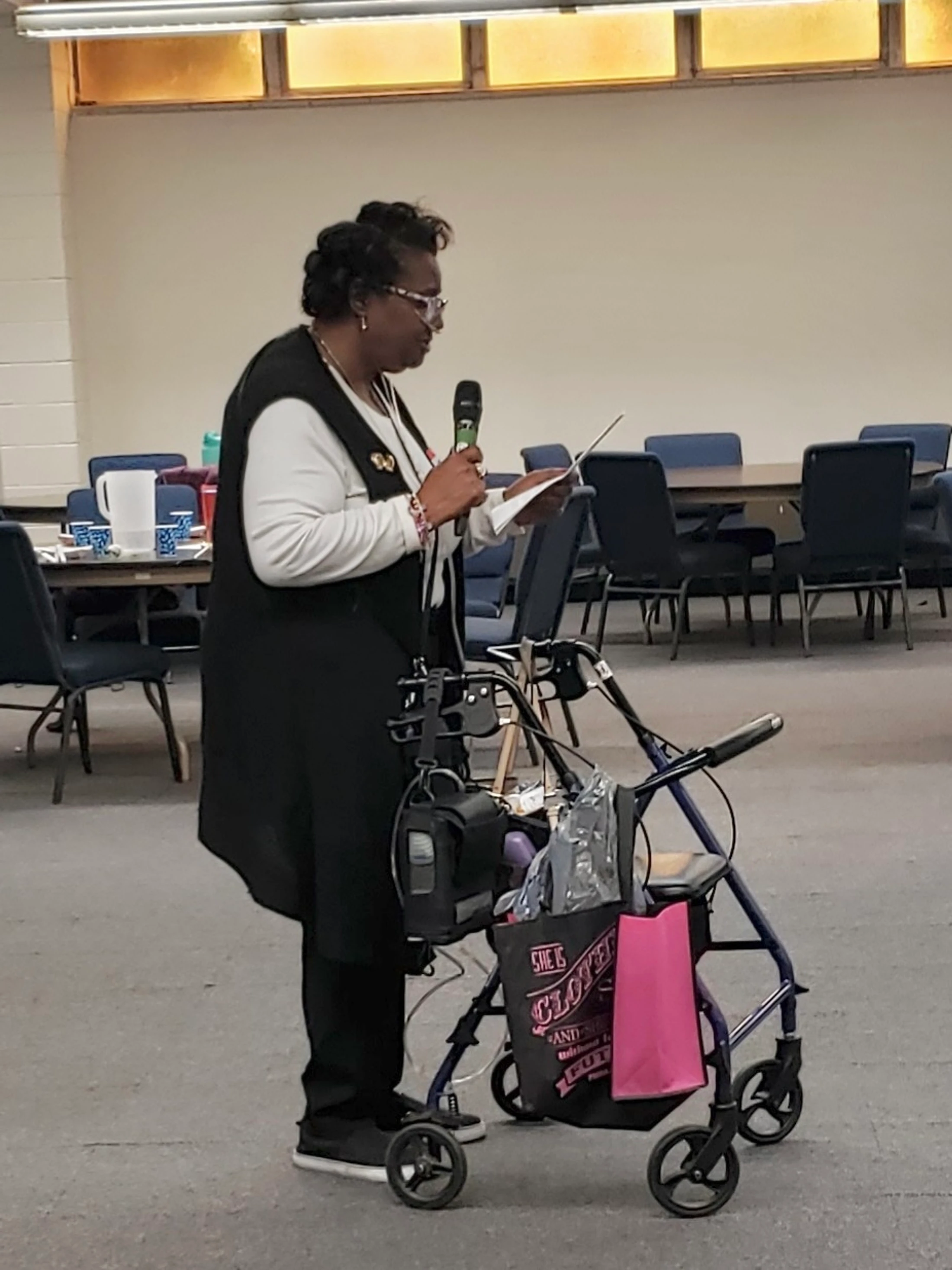 An older Black woman stands next to her purple walker, speaking into a microphone and reading from a paper in a large hall with tables and chairs.
