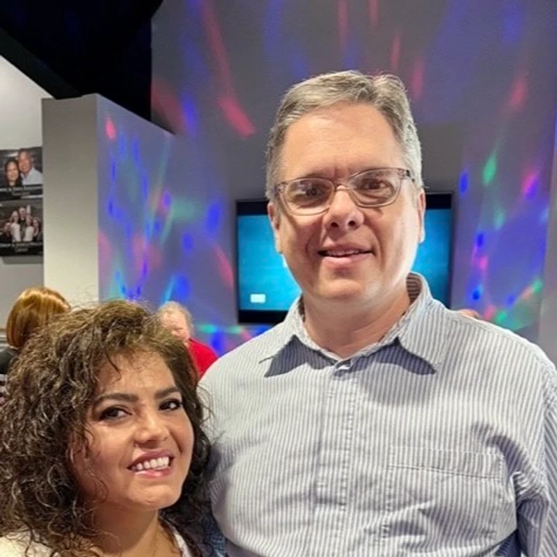 A smiling couple, a woman with curly hair and a man with glasses, pose for a photo in a room with blue and pink disco lights in the background.