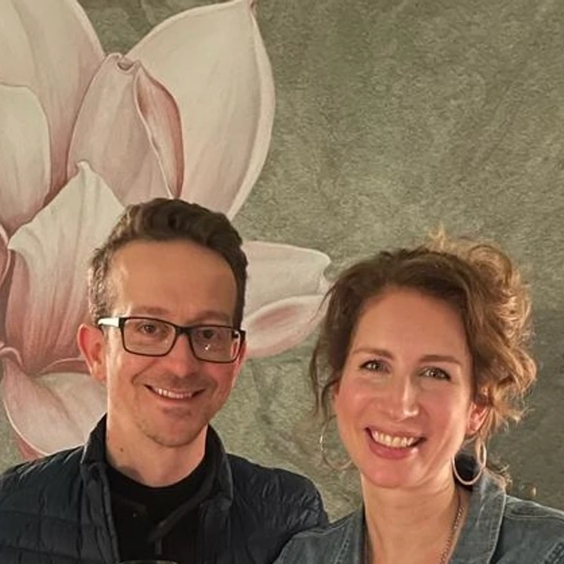 A man with glasses and a woman with curly hair smile for a photo in front of a wall with a large pink and grey floral pattern.