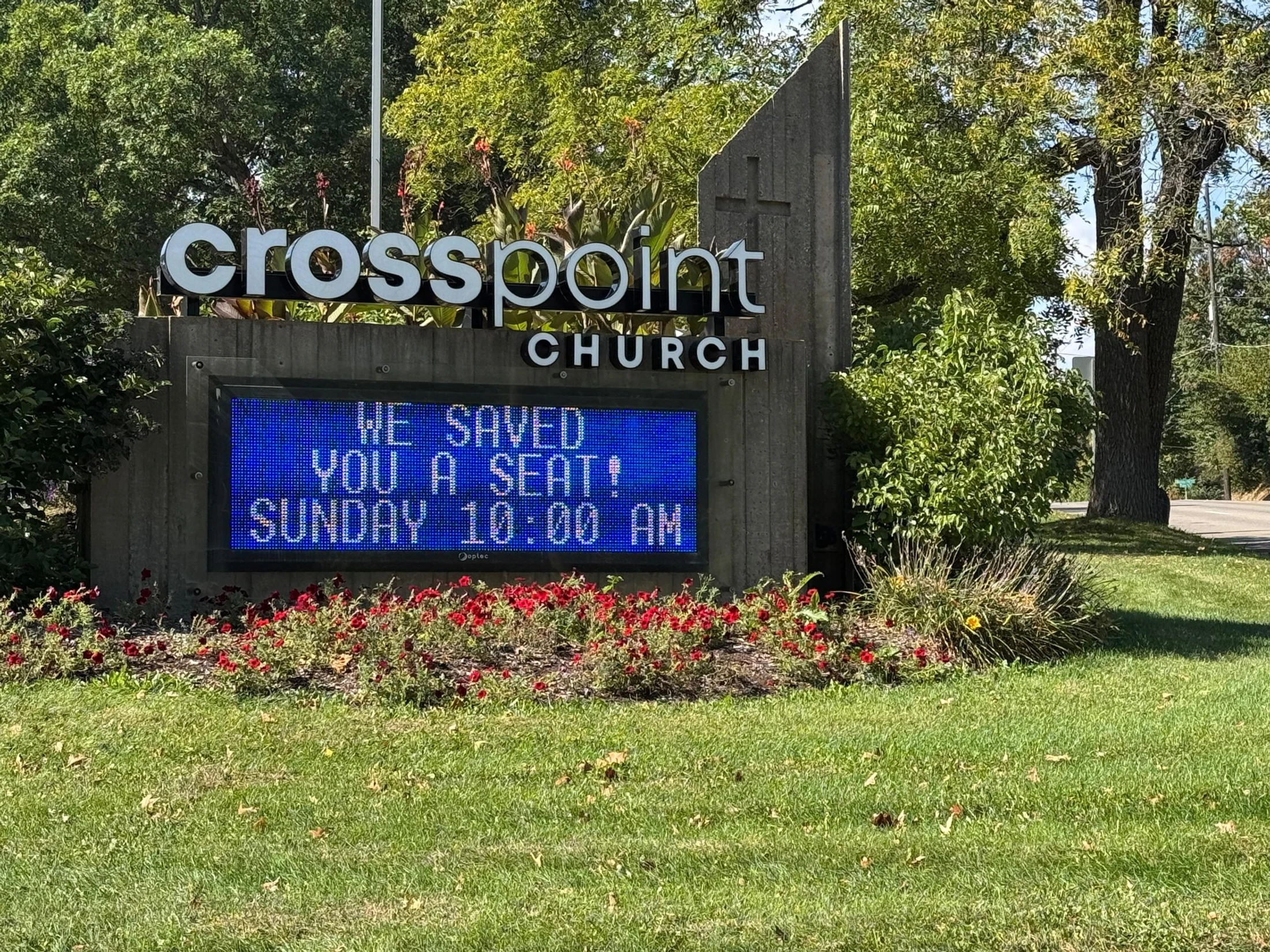 A concrete sign for Crosspoint Church with a blue digital display reading 'WE SAVED YOU A SEAT! SUNDAY 10:00 AM' set on a lawn with red flowers.