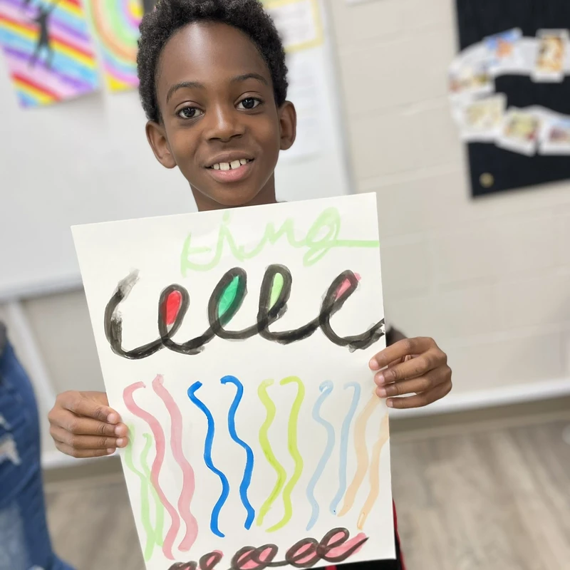 A smiling young Black boy holds up a colorful painting with black swirls and wavy vertical lines that he made in a classroom.