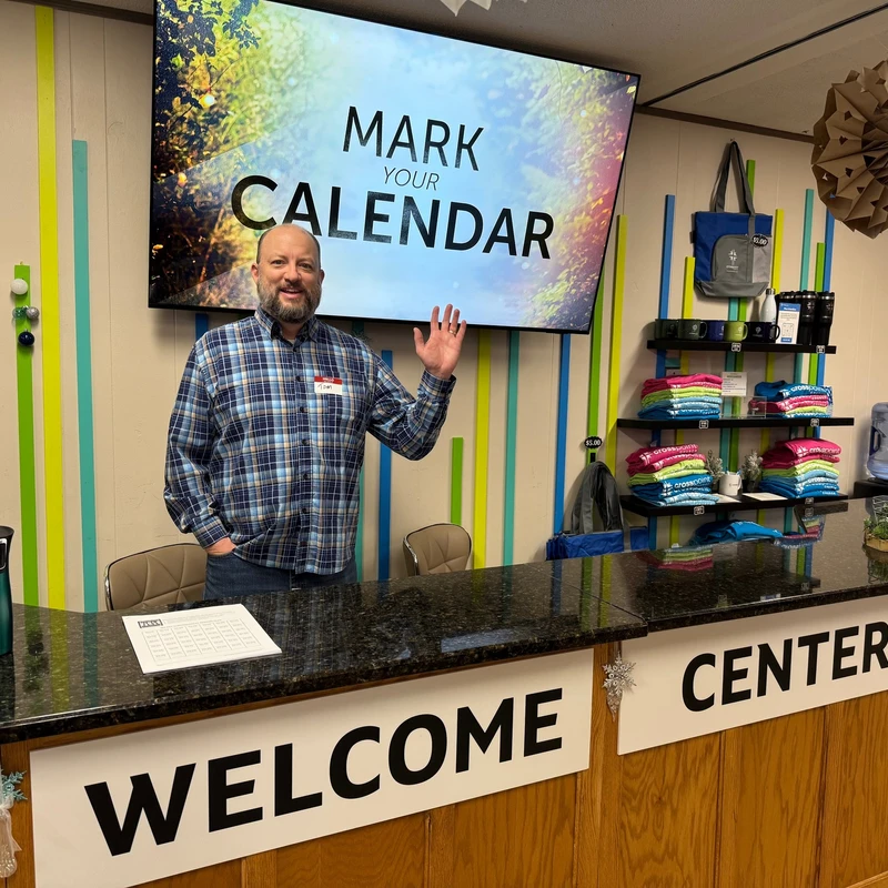 A smiling man in a plaid shirt waves from behind a welcome center desk. A large screen behind him reads, 'Mark Your Calendar'.