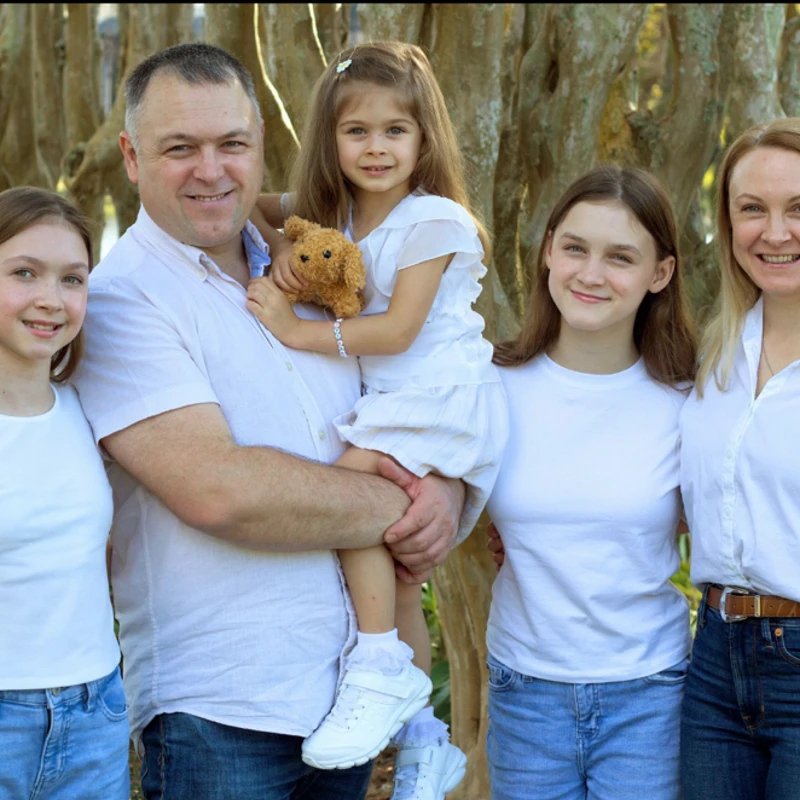 A family of five, parents and three daughters, pose for a photo outdoors. They are all smiling and wearing white shirts and blue jeans.