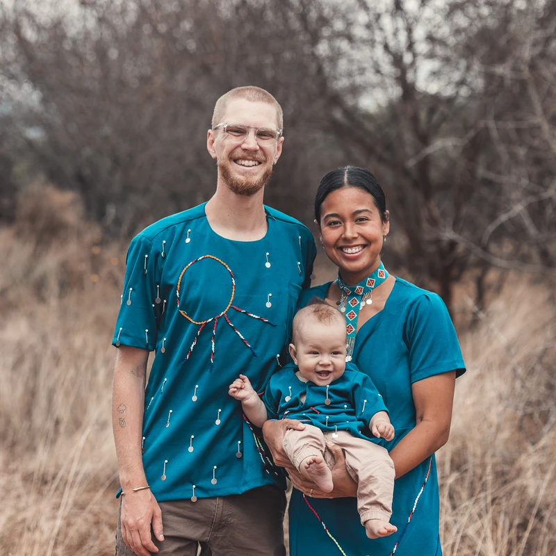 A smiling man, woman, and baby pose outdoors in a grassy field. They are all wearing matching teal-colored outfits with colorful beaded decorations.