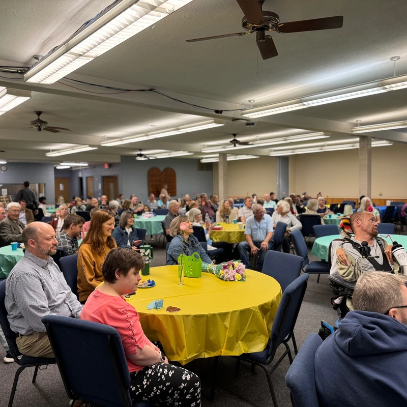 A diverse crowd of people seated at round tables with colorful tablecloths in a brightly lit hall, attentively watching a presentation or speaker.