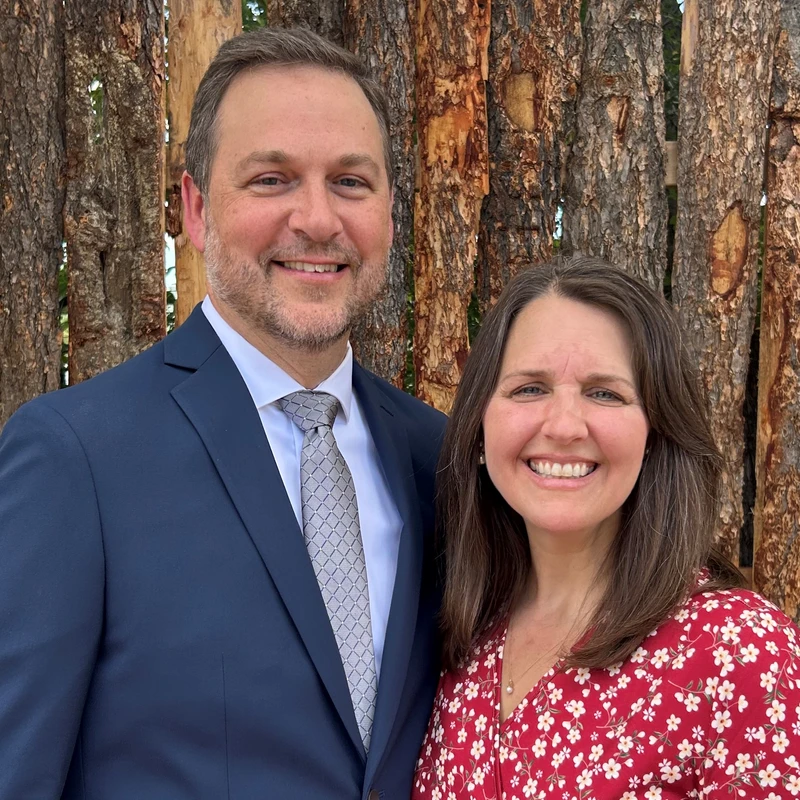 A man in a blue suit and a woman in a red floral dress stand together smiling in front of a wall made of vertical tree trunks.