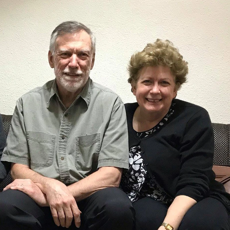 A portrait of an older man with a grey beard and a woman with short curly hair sitting side-by-side on a couch, both smiling warmly at the camera.