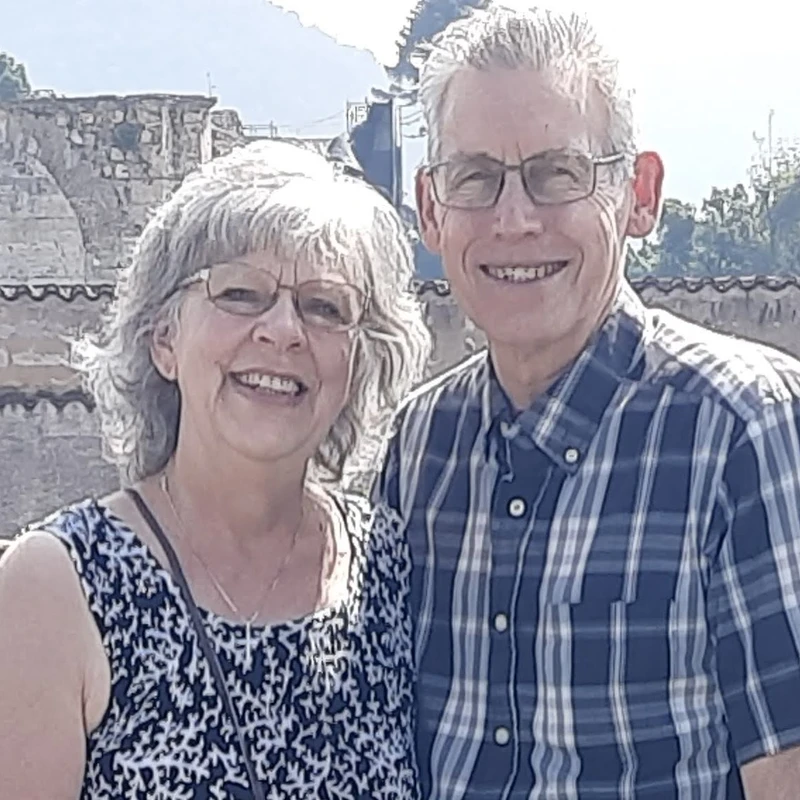 An older man and woman with grey hair and glasses smile for the camera. They stand outside with old stone buildings and tiled roofs in the background.