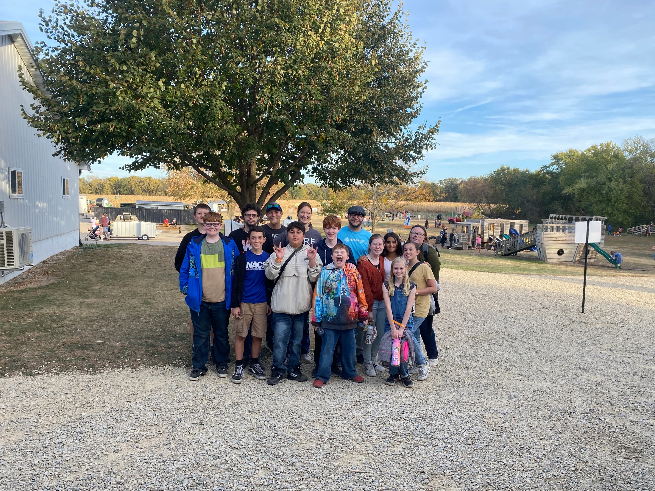 A group of about fifteen children and adults posing together for a photo under a large tree on a gravel lot, with a playground and farm in the background.