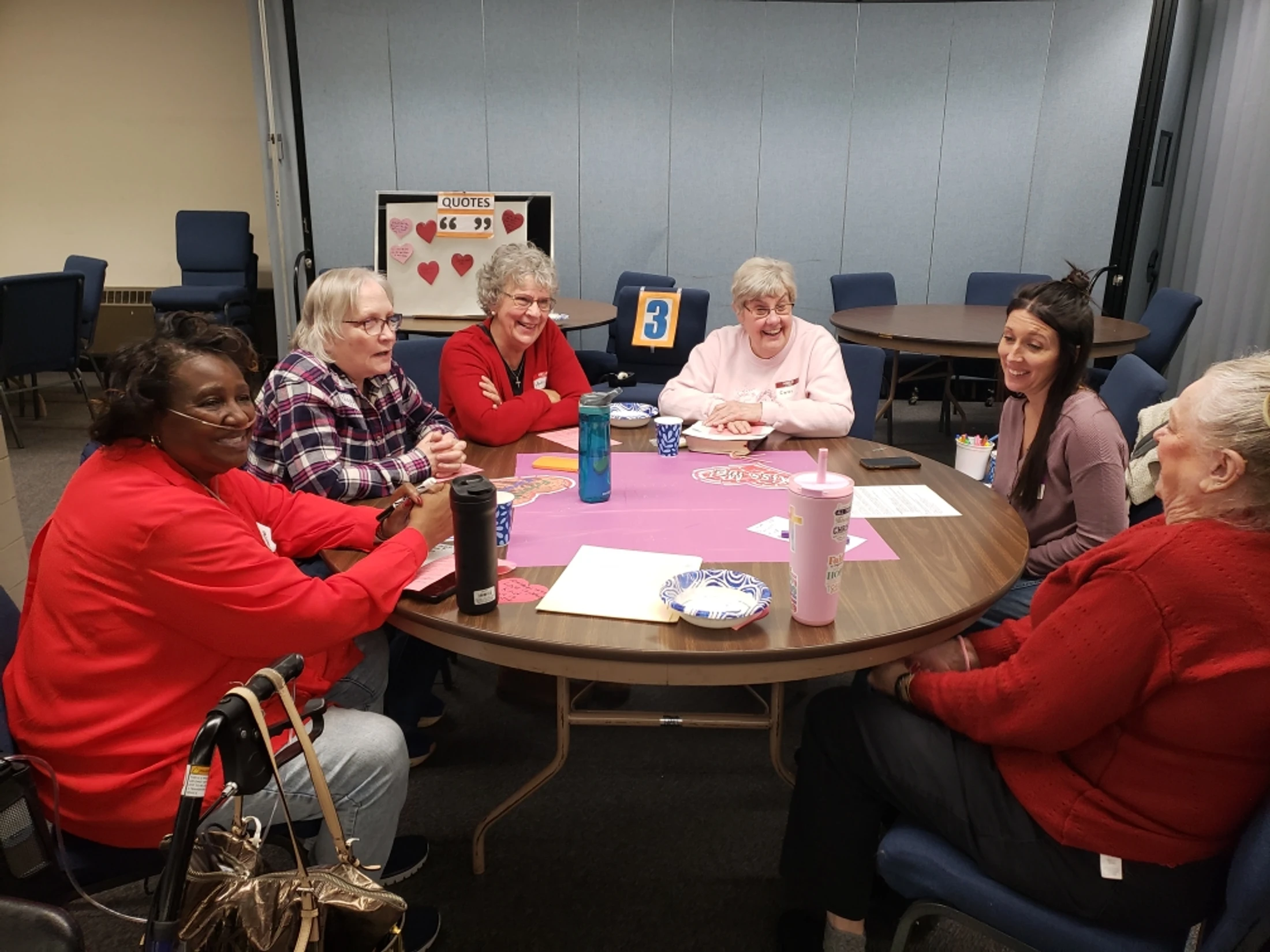 A diverse group of six women sit smiling and laughing together around a circular table in a community room, engaged in a lively conversation.