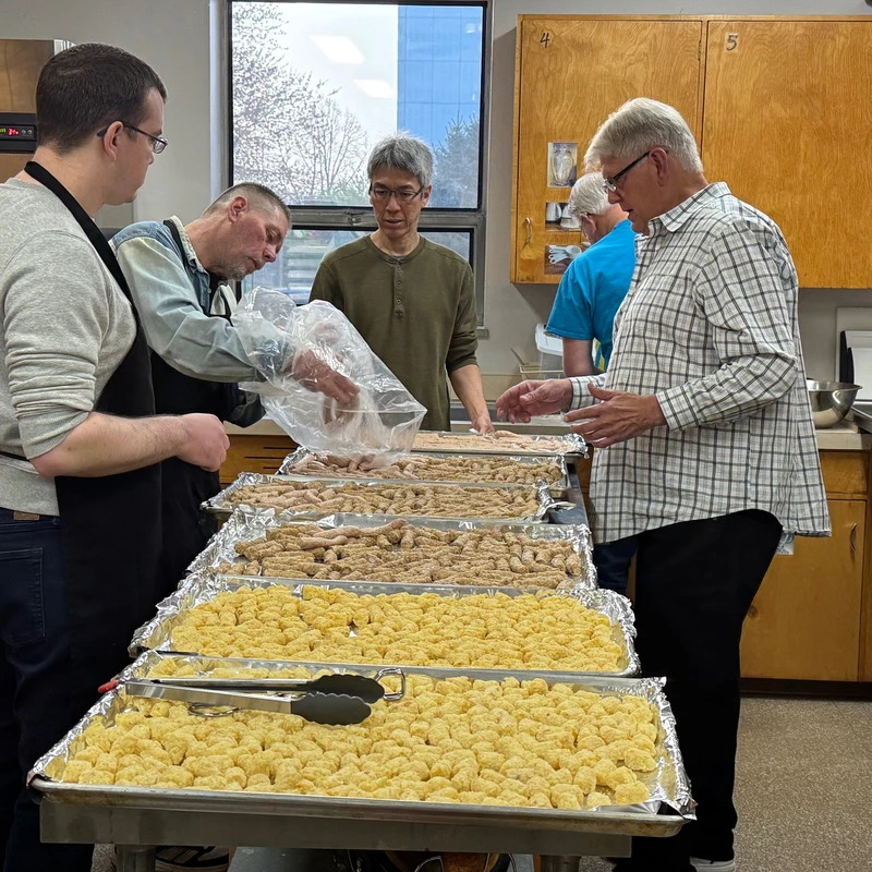 Five men stand in a large kitchen preparing food. They are gathered around a counter with large trays of tater tots and sausages.