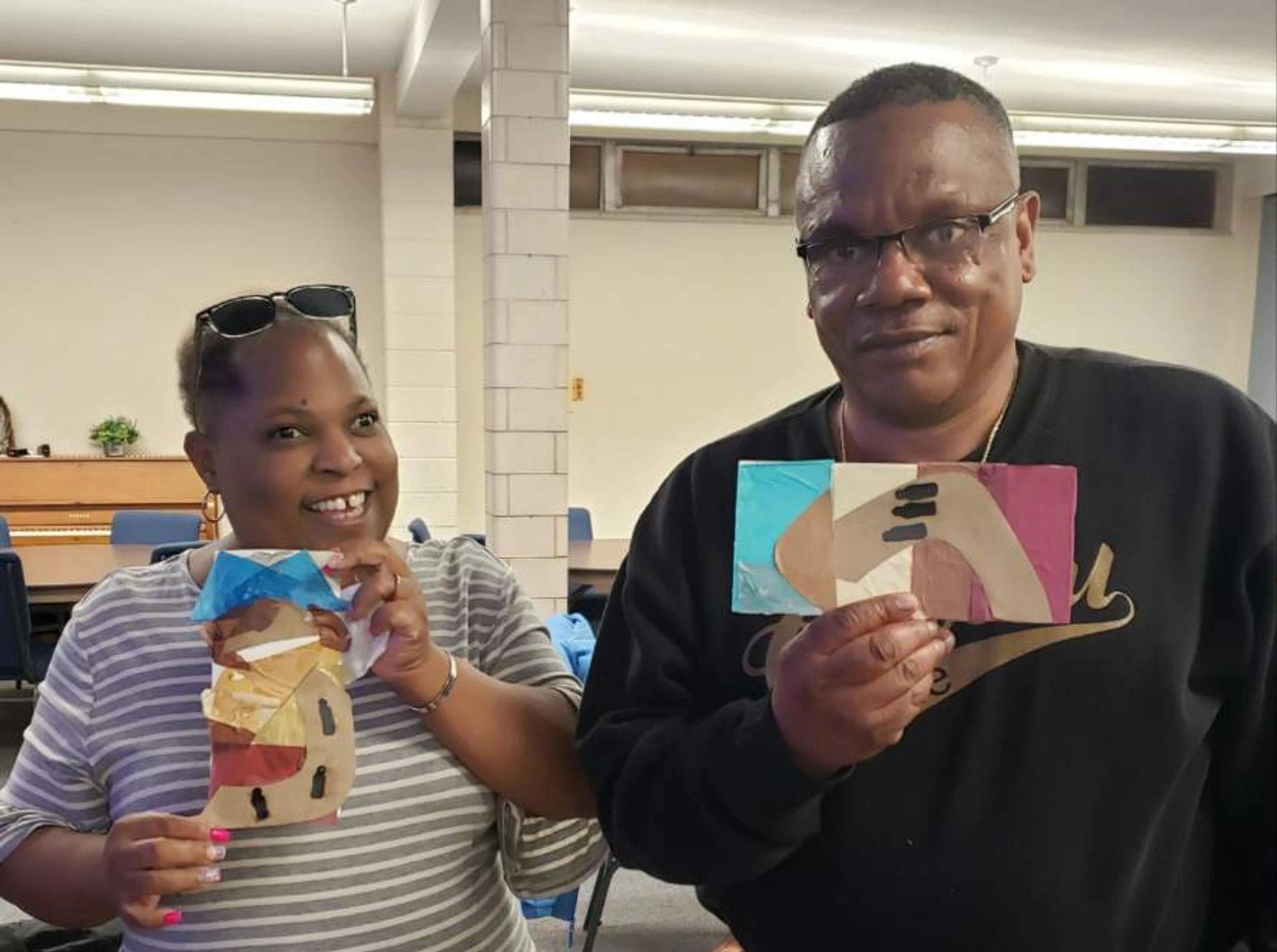 A smiling woman in a striped shirt and a man in a black sweatshirt hold up colorful paper collages they created during an indoor art class.