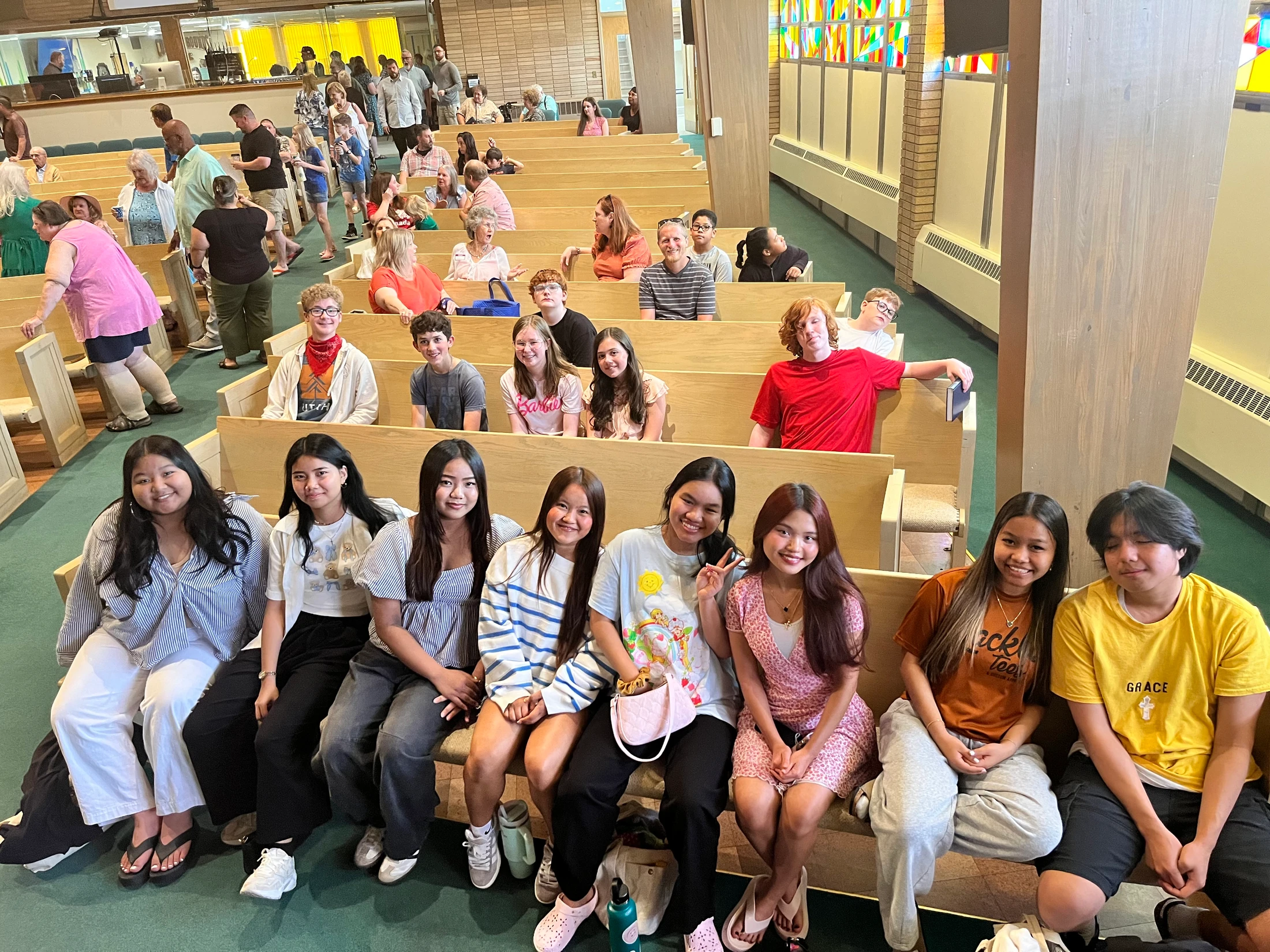 A diverse group of young people smiles for a photo while sitting on a wooden pew inside a church. Other congregation members are in the background.
