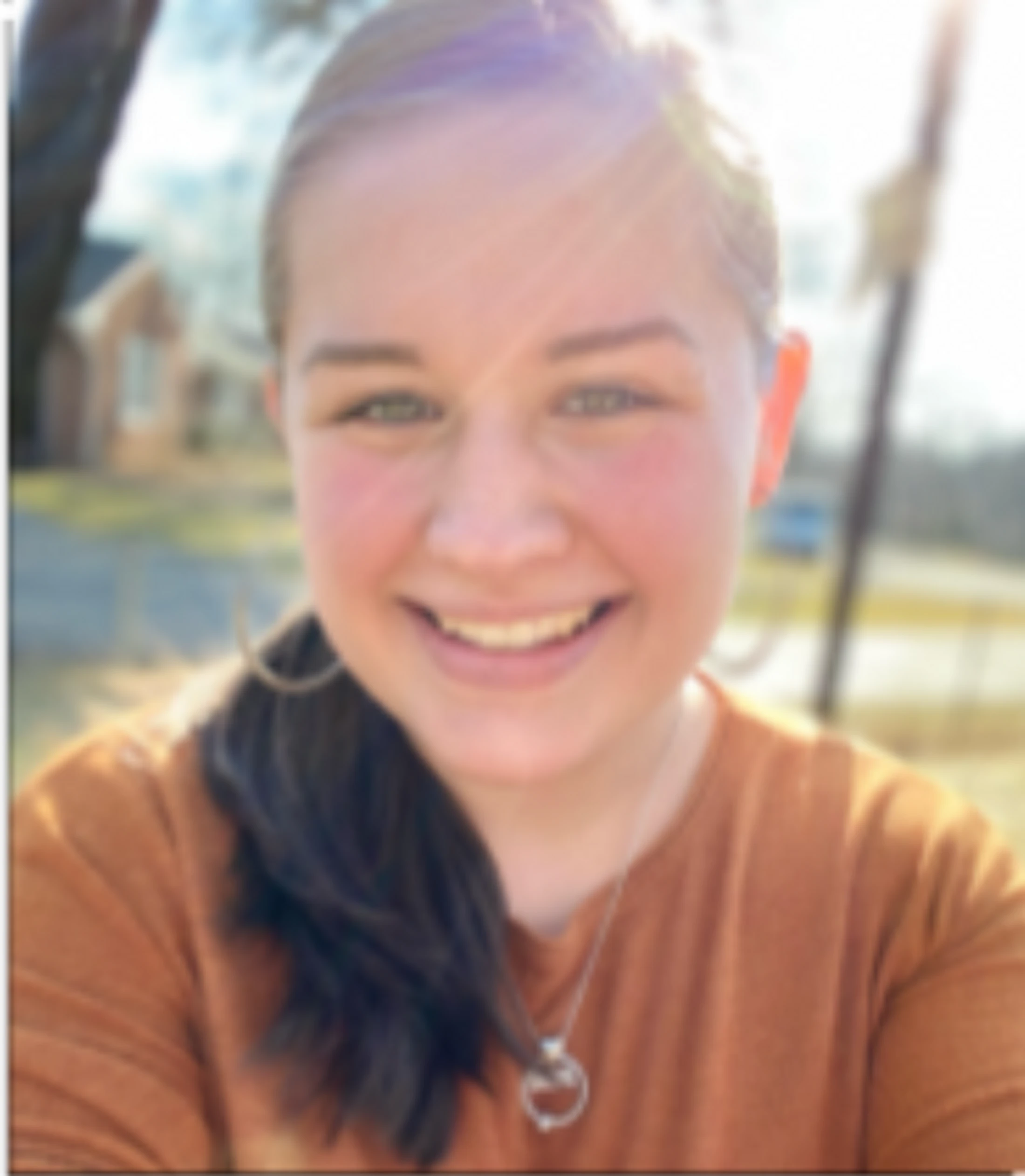 A close-up headshot of a young woman with brown hair pulled back, smiling at the camera. She is outdoors on a sunny day wearing an orange shirt.