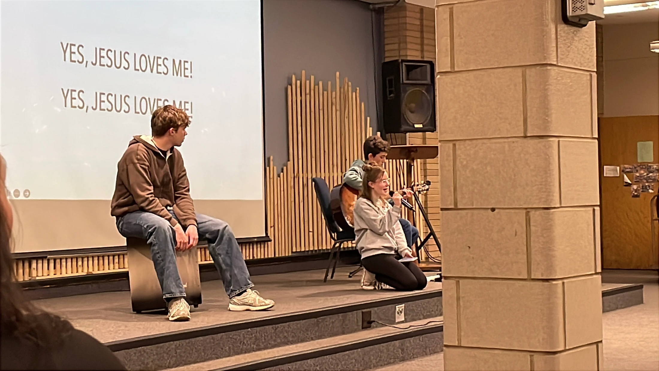 Three young people on a stage during a worship service. A girl kneels singing, a boy plays guitar, and a screen says "YES, JESUS LOVES ME!".