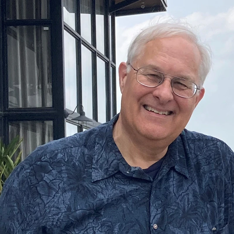 A smiling older man with white hair and glasses wearing a blue patterned shirt, standing on a rooftop or balcony with a cityscape in the background.