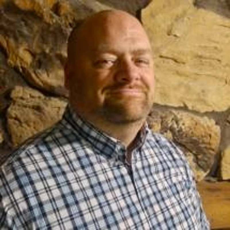 A smiling, bald man with a short beard, wearing a blue and white plaid shirt, poses for a headshot in front of a stone wall.