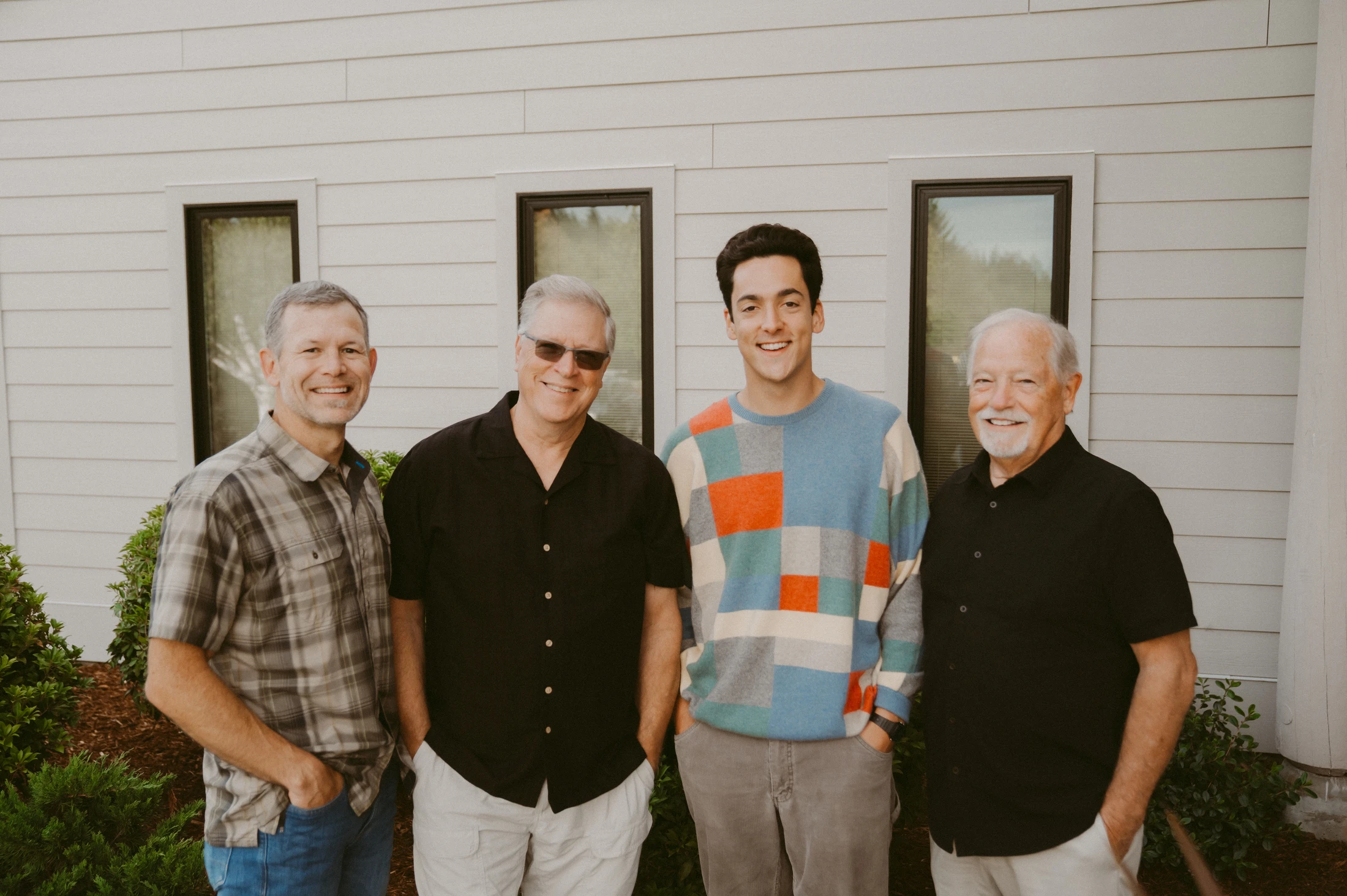 Four men of varying ages stand side-by-side and smile for a photo outdoors in front of a light-colored building.