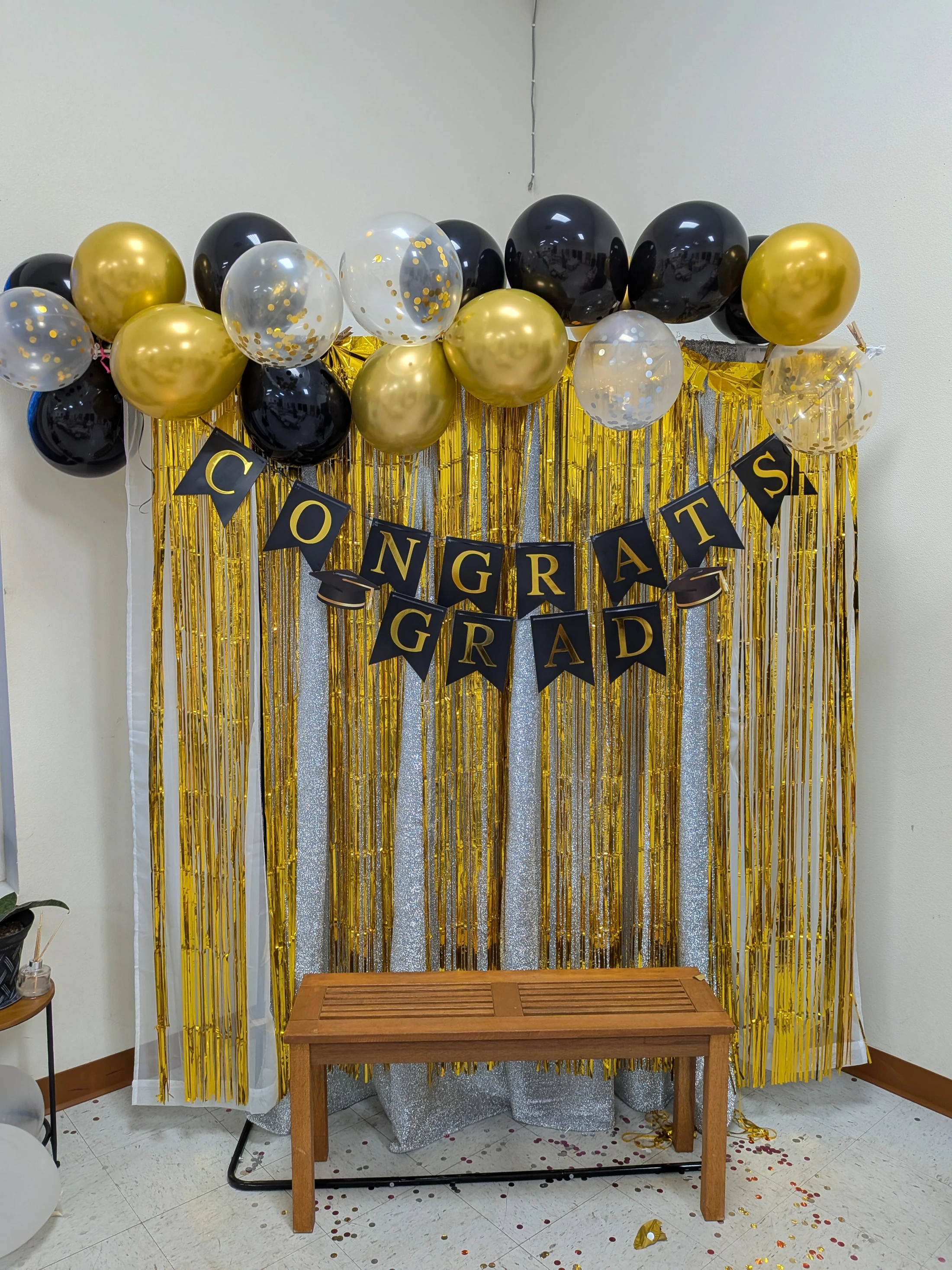 A photo booth for a graduation party with a gold and silver tinsel backdrop, a 'Congrats Grad' banner, an arch of black and gold balloons, and a wooden bench.