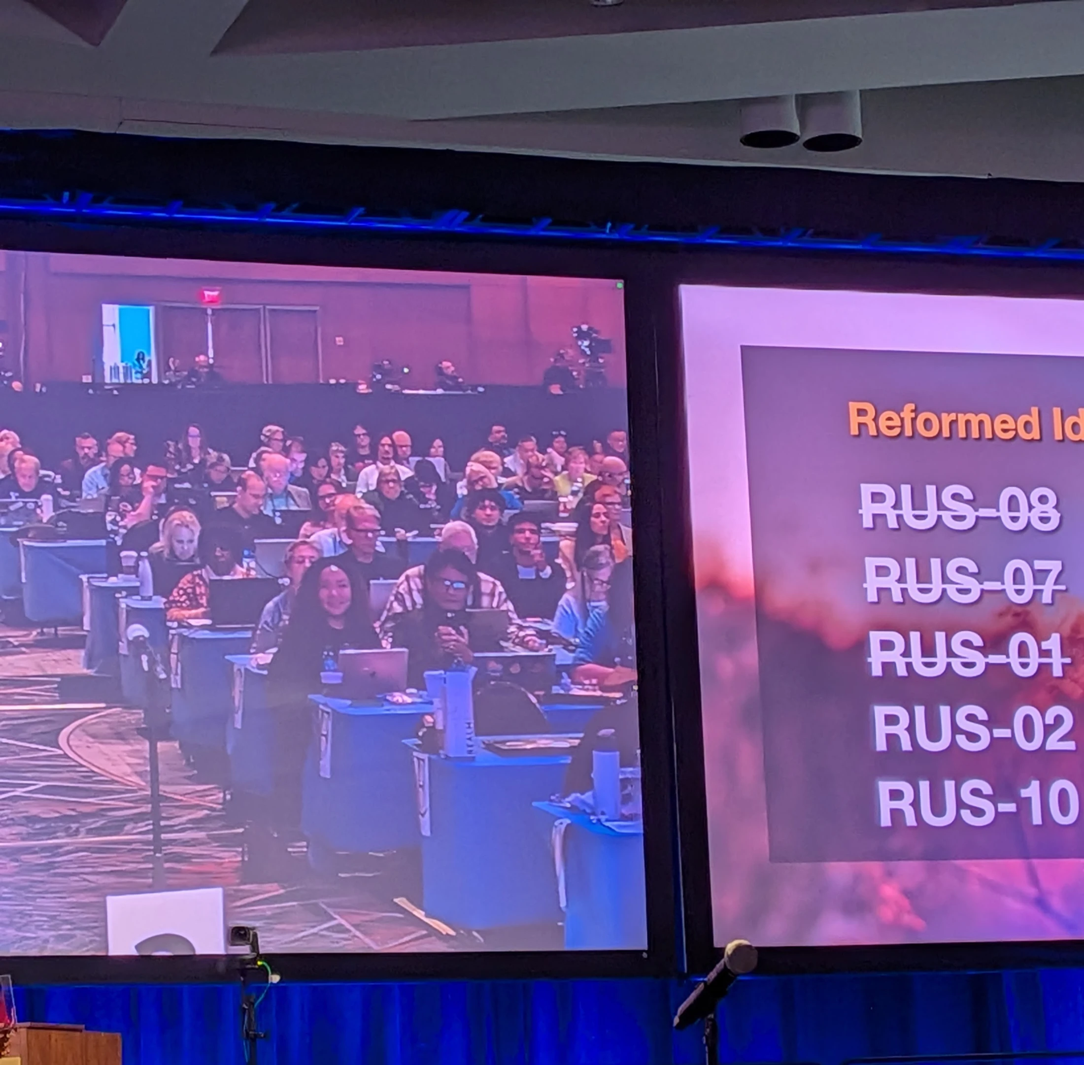 Two large screens at a conference. One shows the audience at their tables with laptops, the other displays a presentation slide with a list of items.