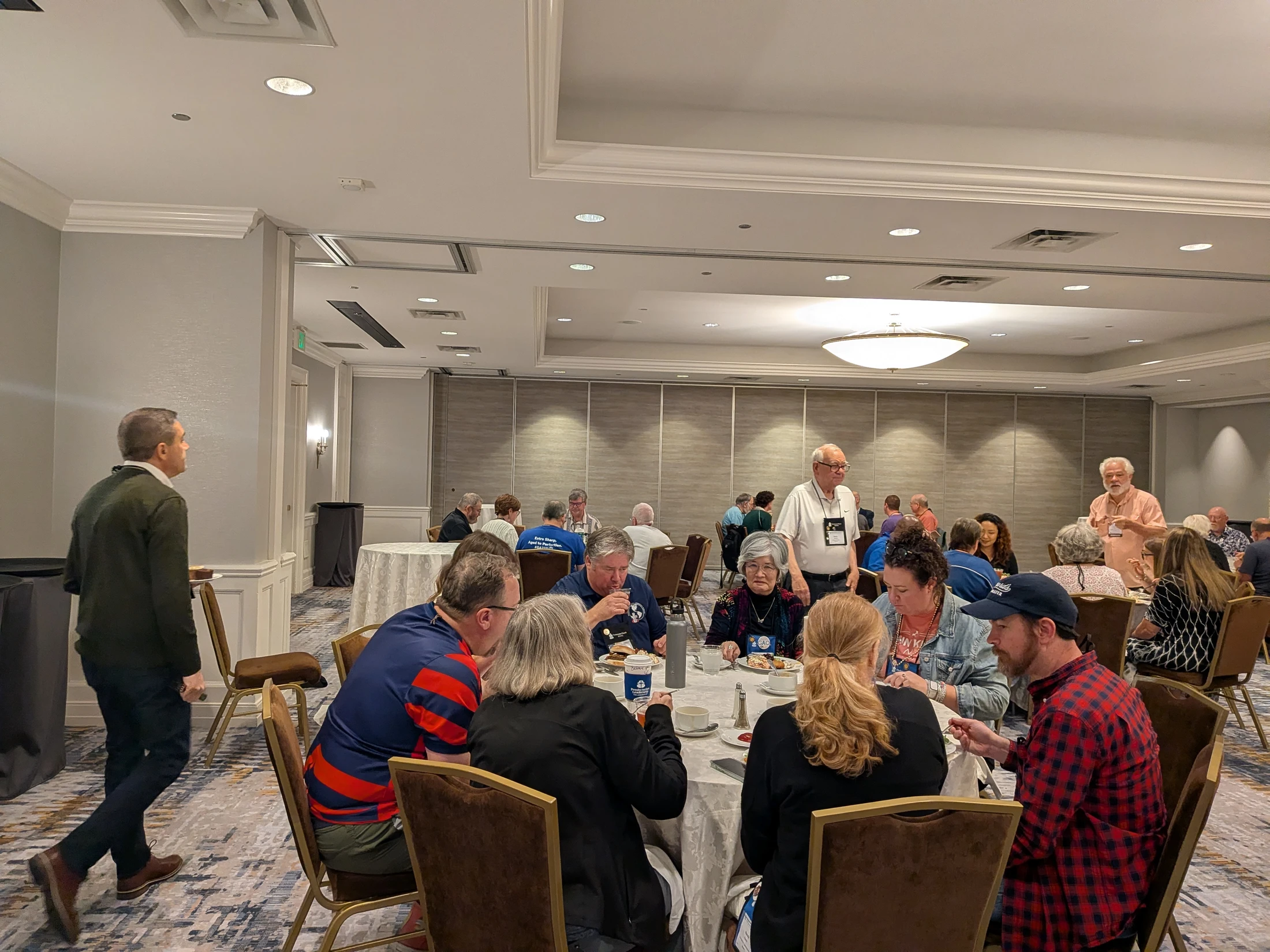 A large group of people eating and socializing at round tables in a spacious hotel conference room during an event.