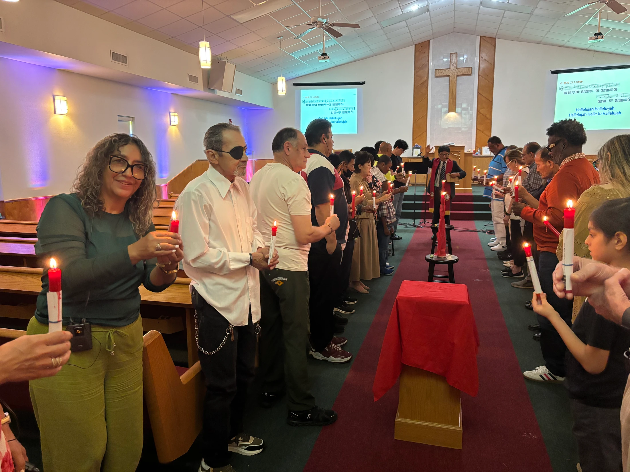 A diverse group of people stand in two lines in a church aisle, each holding a lit red candle during a worship service with song lyrics on a screen.