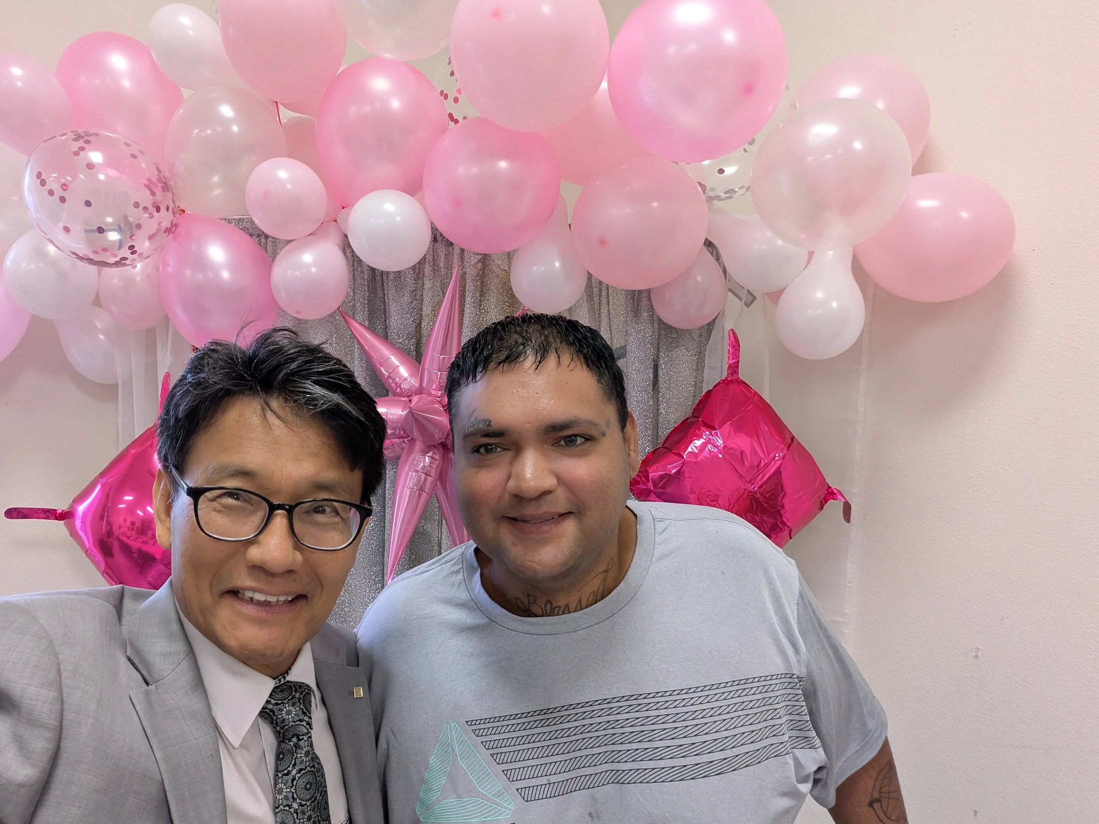 Two smiling men take a selfie in front of a celebratory backdrop of pink and white balloons and a silver curtain. One man wears a suit, the other a t-shirt.