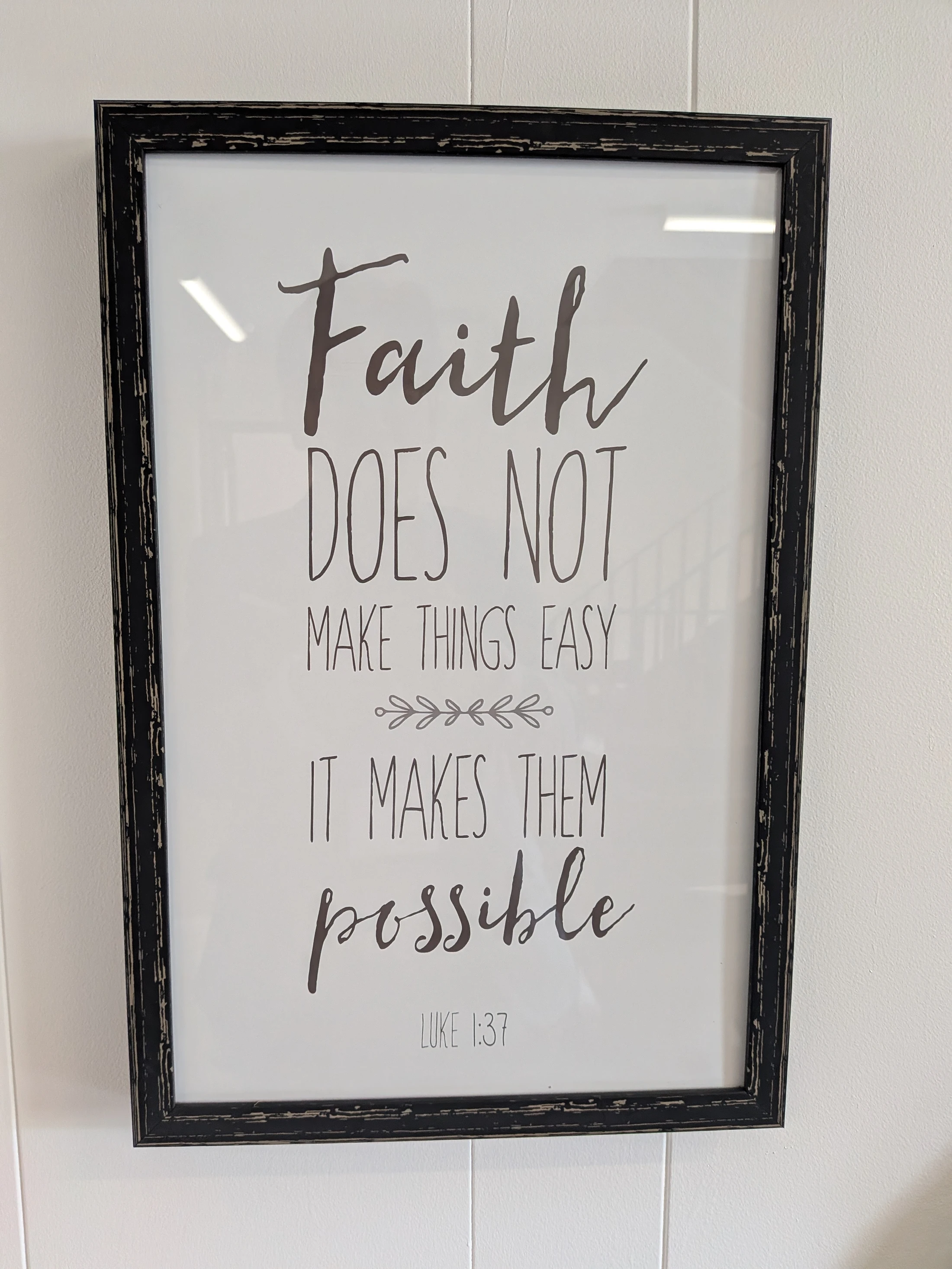 A framed print with a distressed black frame hangs on a white wall. It reads, "Faith does not make things easy it makes them possible," with "Luke 1:37" below.