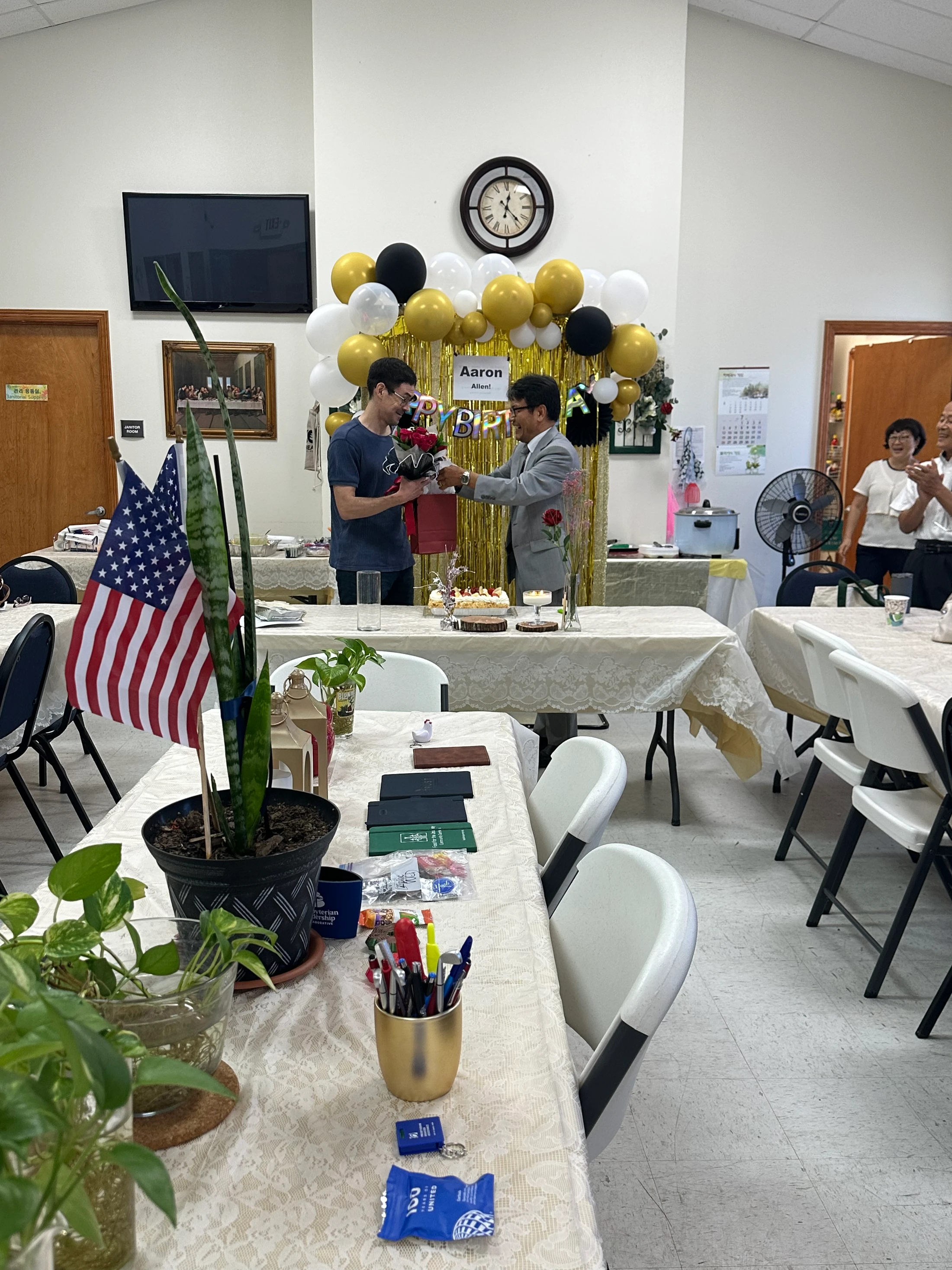A man in a suit gives a bouquet of red roses to another man at his birthday party, celebrated with balloons, a banner, and cake in a decorated room.