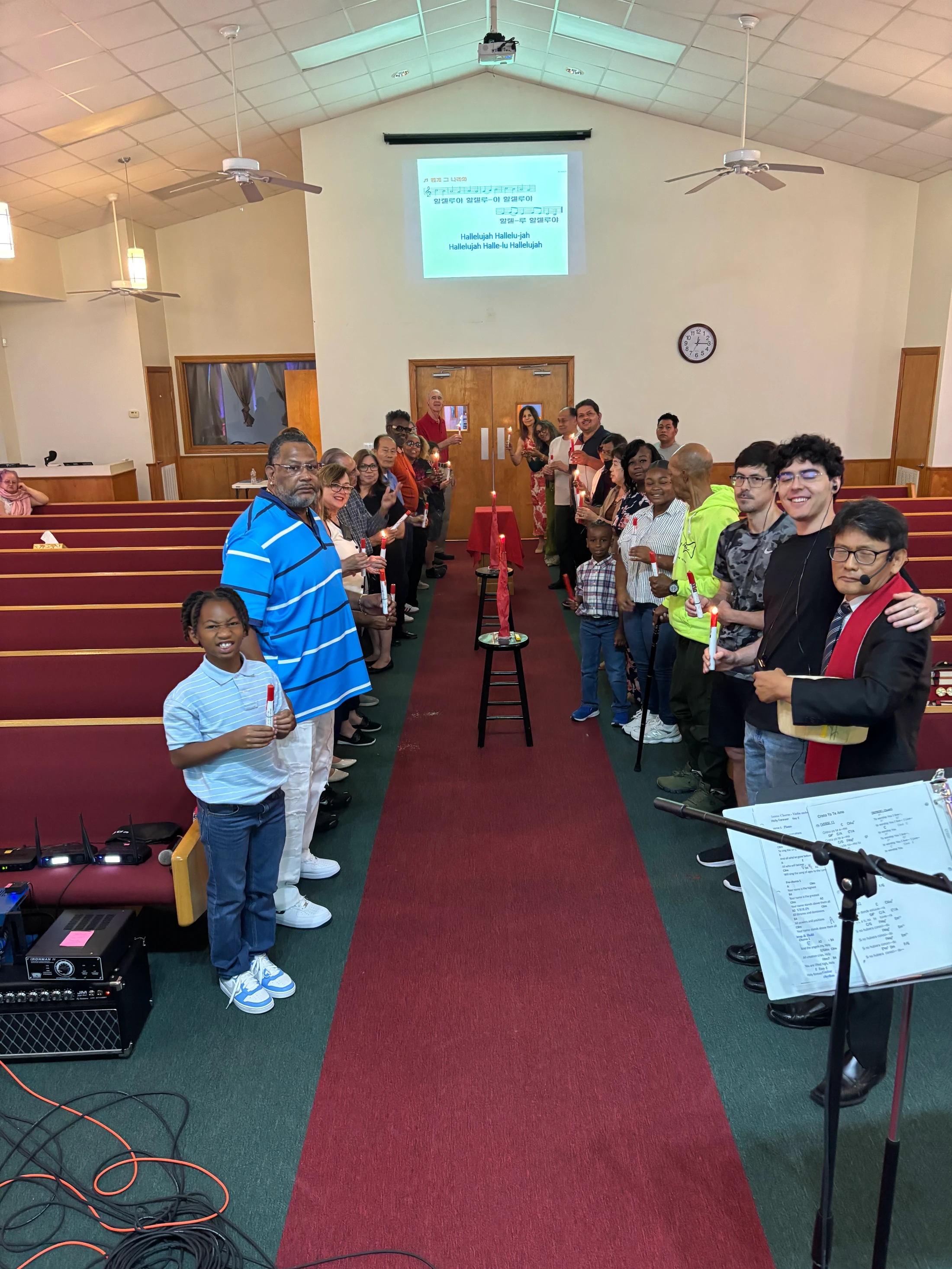 A diverse congregation stands in a church aisle holding candles during a ceremony. A screen in the background displays lyrics in Korean and English.