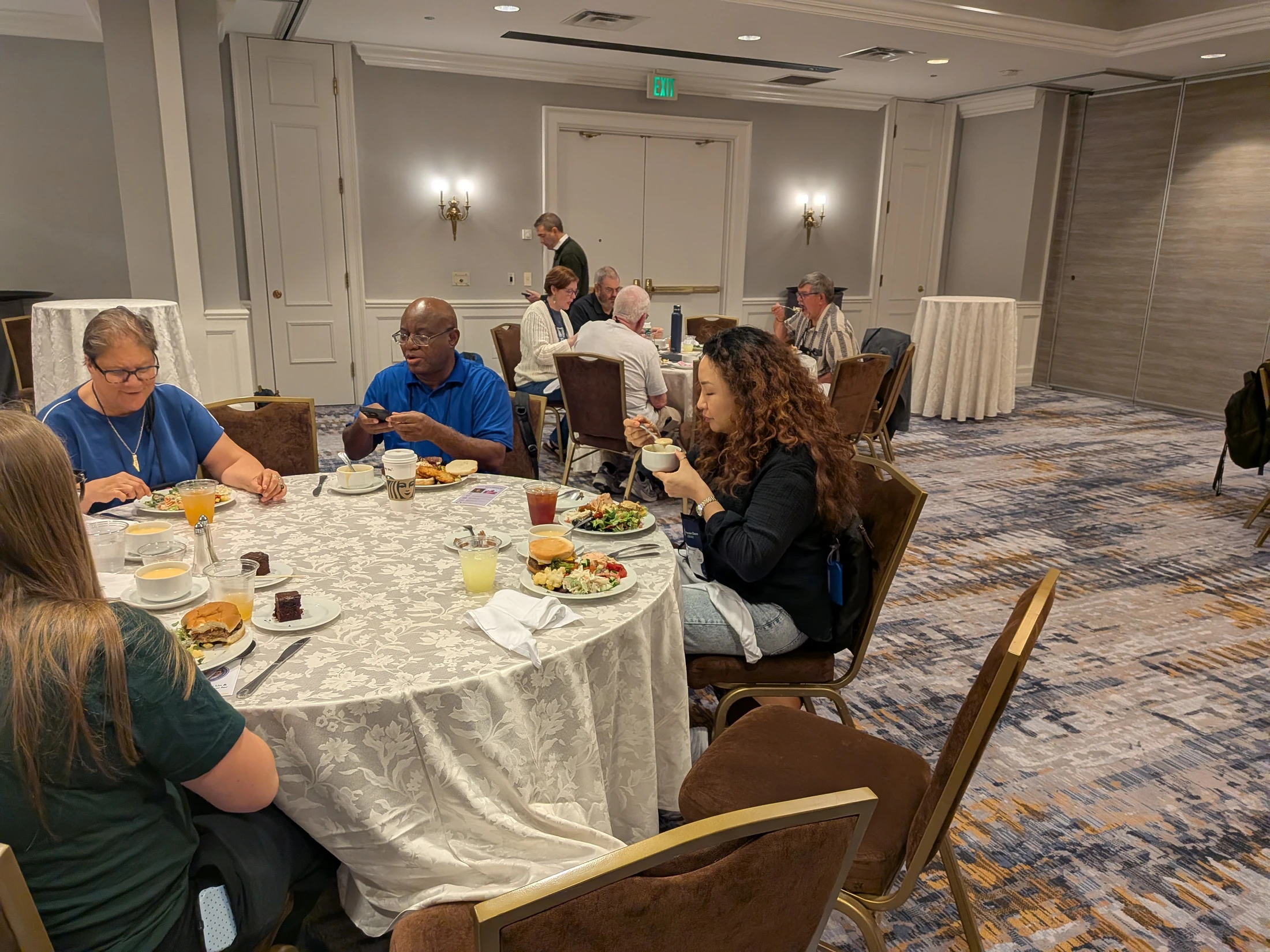 A diverse group of people sitting at round tables in a conference room, eating lunch. The tables are covered with white, patterned tablecloths.