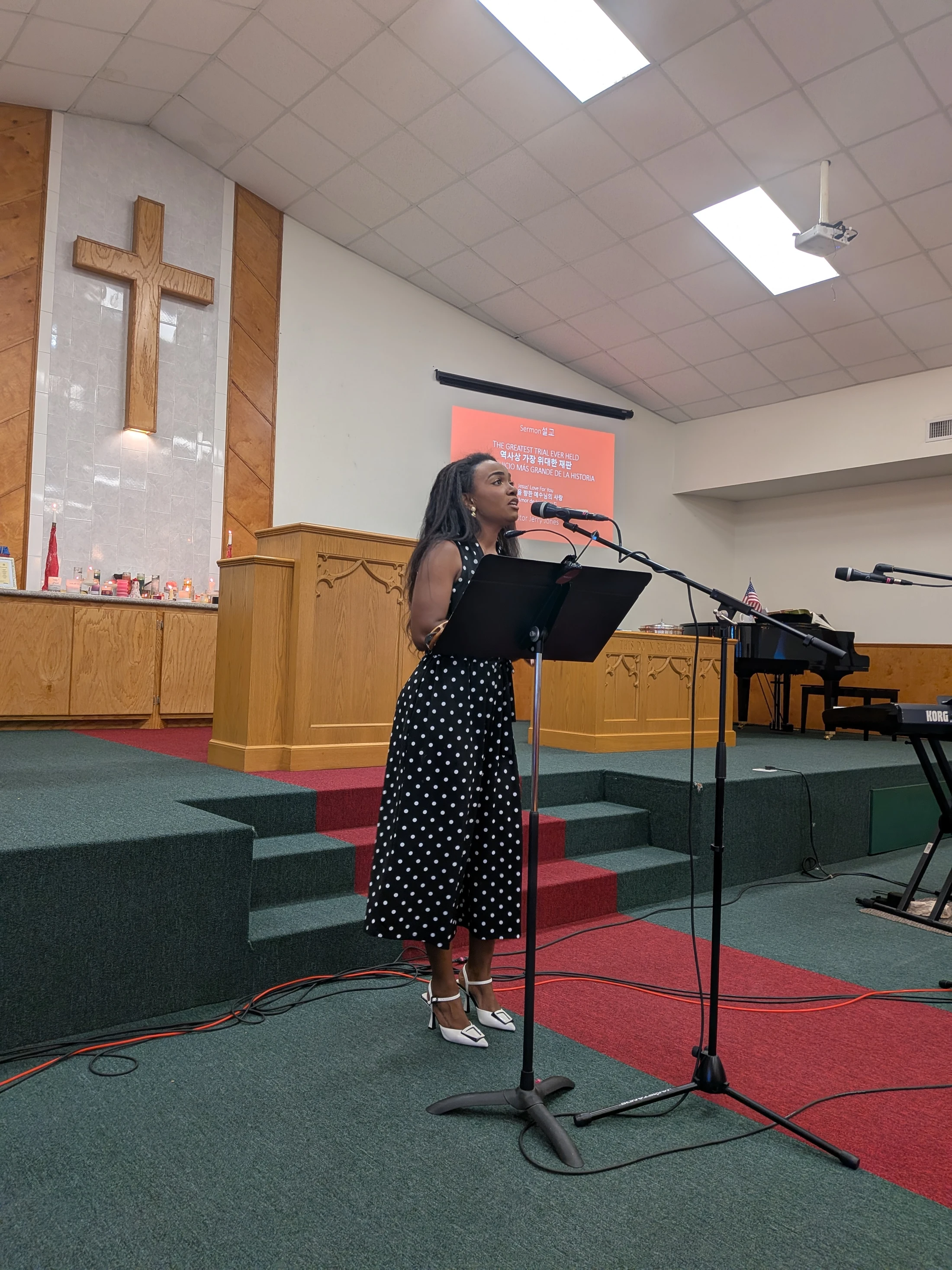 A Black woman in a black and white polka-dot dress stands at a podium on a church stage, speaking into a microphone. A wooden cross is on the wall behind her.