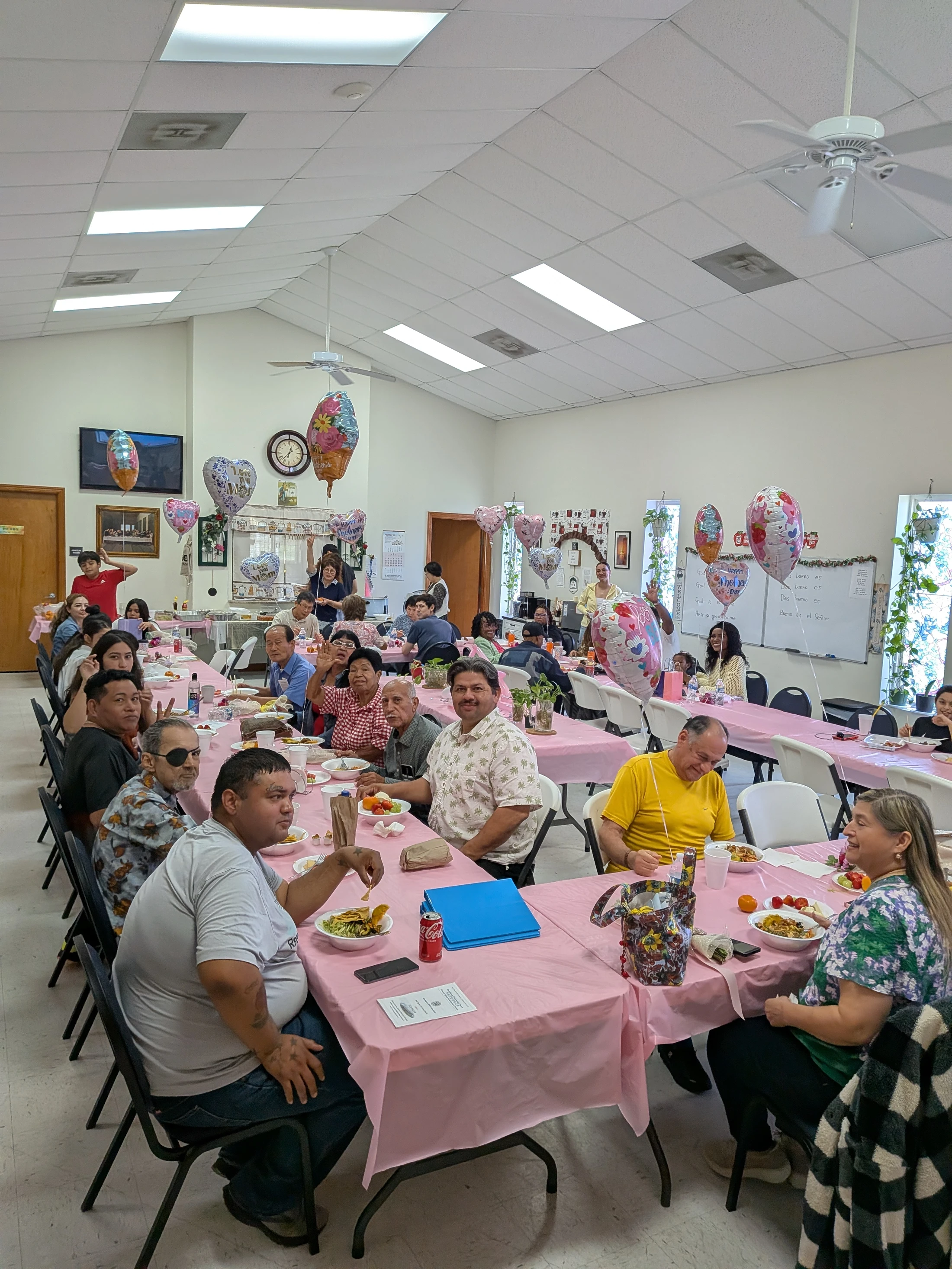 A large, diverse group of people celebrating at long tables with pink tablecloths and festive balloons in a brightly lit community hall.