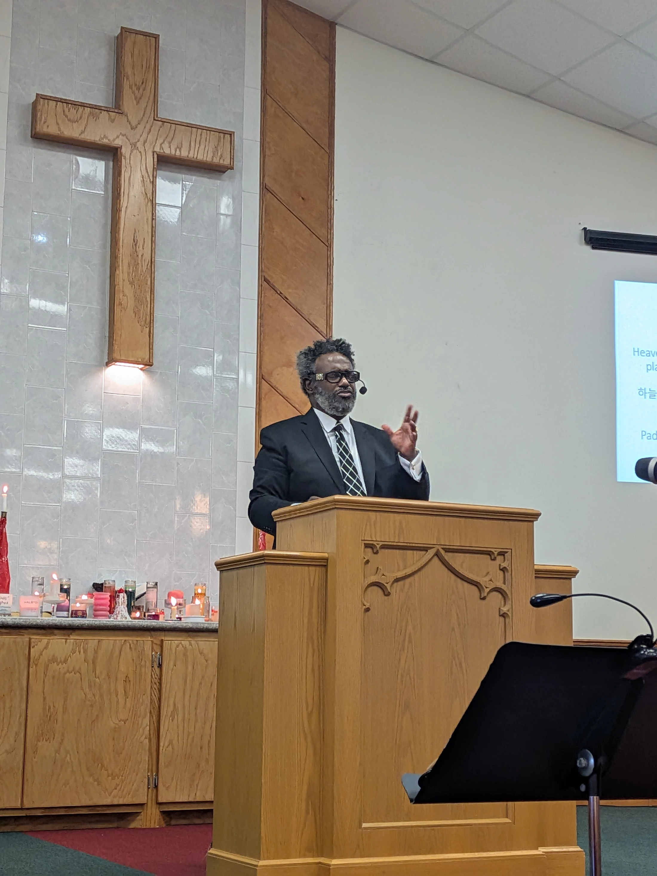 A Black pastor in a suit and tie stands behind a wooden pulpit, giving a sermon. Behind him, a large wooden cross hangs on a tiled wall in the church.