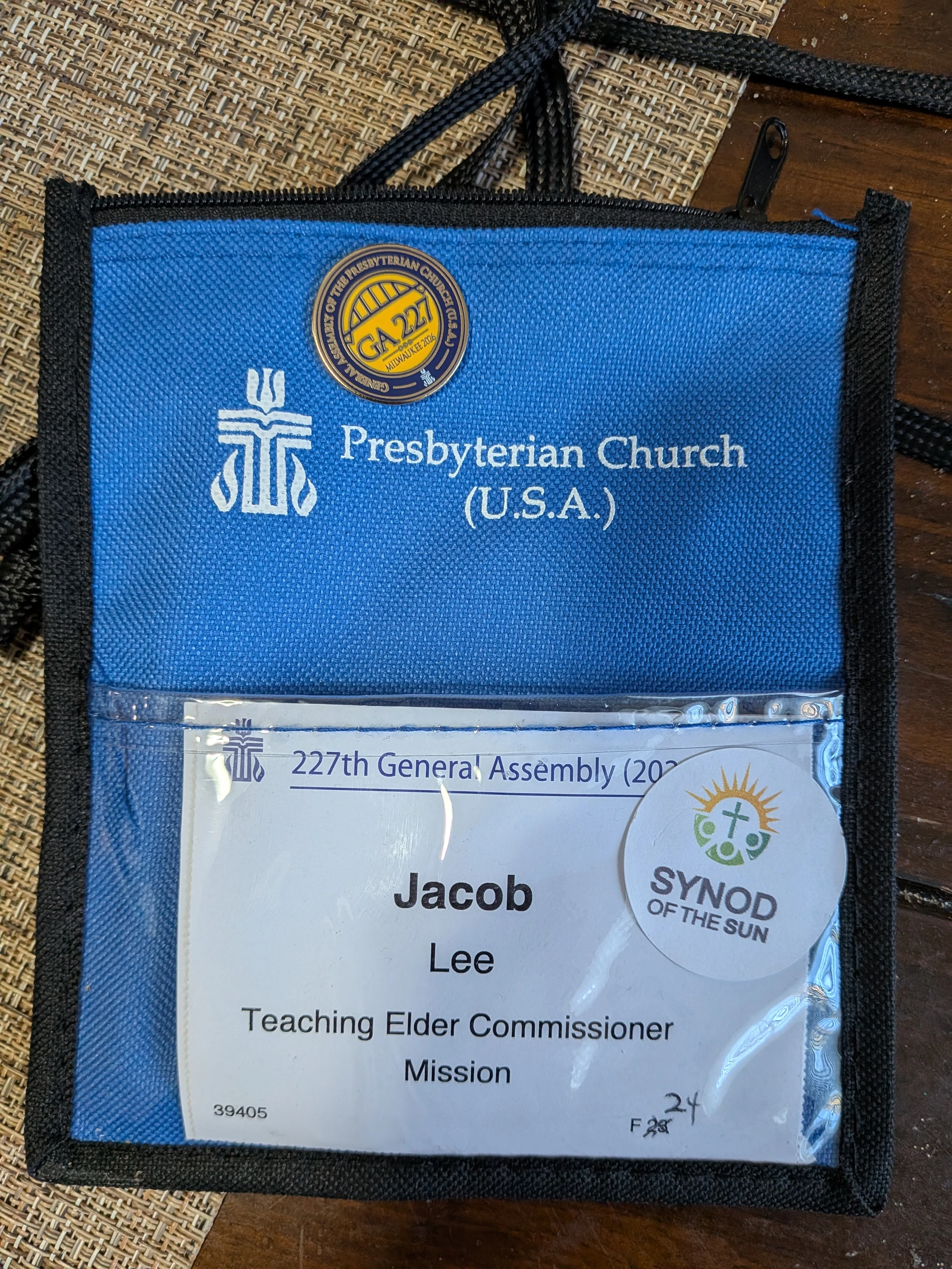 A blue lanyard pouch for the Presbyterian Church (U.S.A.) holding a name badge for the 227th General Assembly for Jacob Lee, Teaching Elder Commissioner.