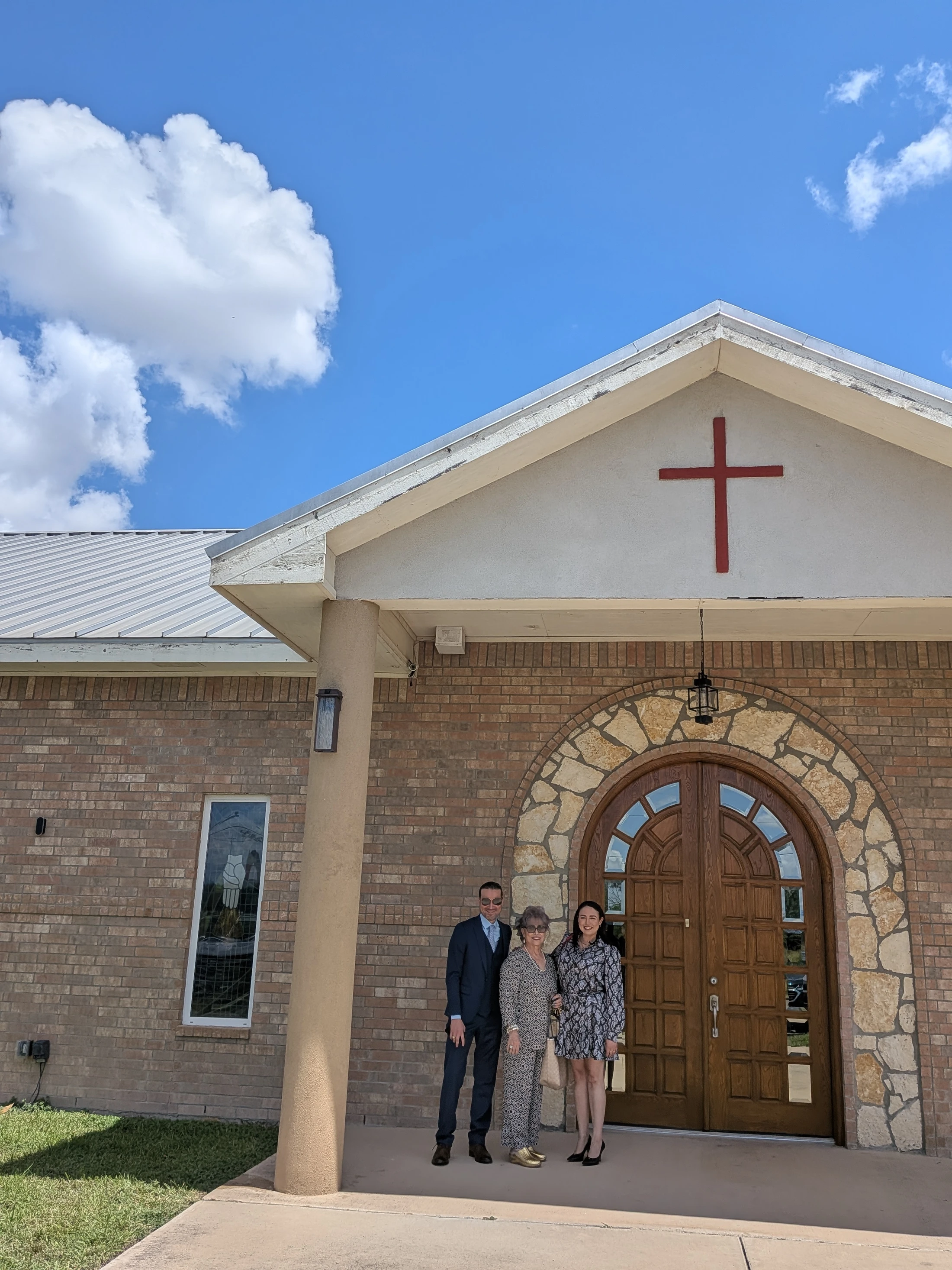 A man in a suit and two women in dresses smile for a picture in front of the arched wooden doors of a brick church on a sunny day.