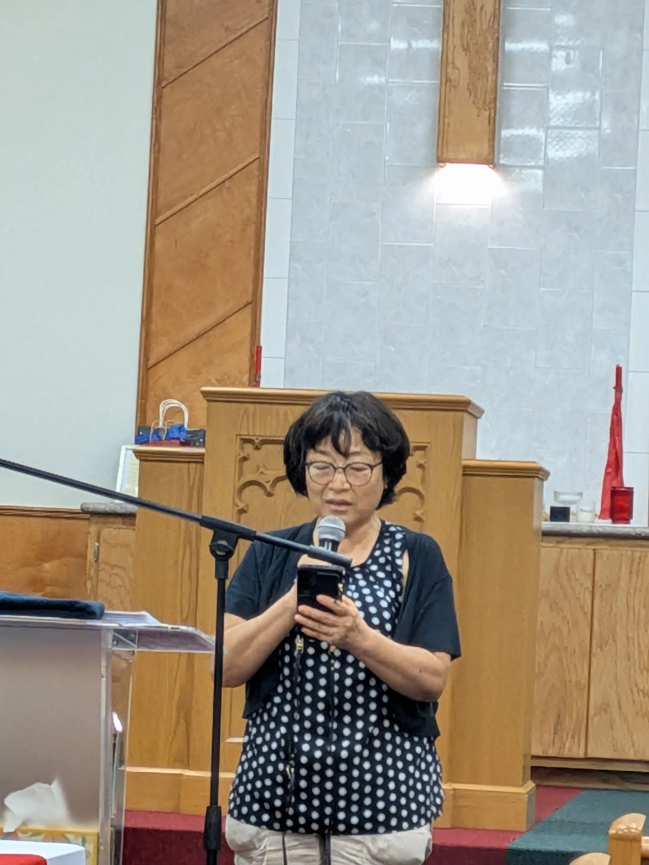 An East Asian woman with short dark hair and glasses stands at a wooden lectern, reading from a smartphone into a microphone during a church service.