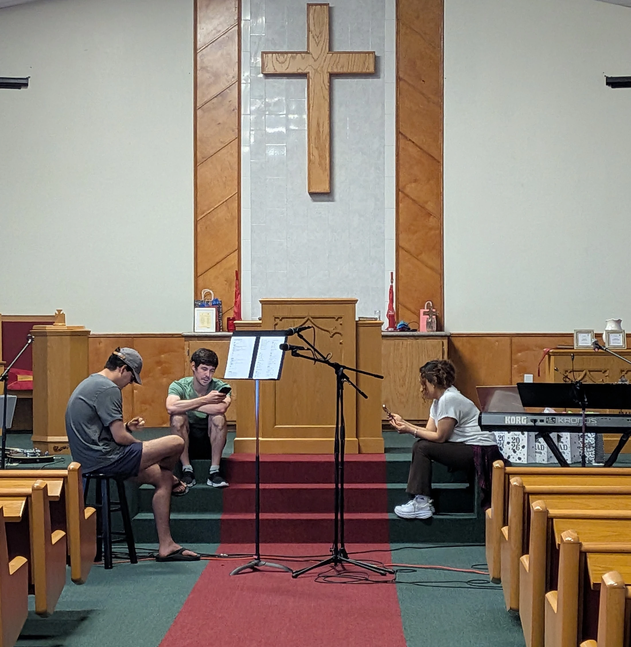 Three young adults sit on a church stage with musical instruments, looking at their phones under a large wooden cross on the wall.