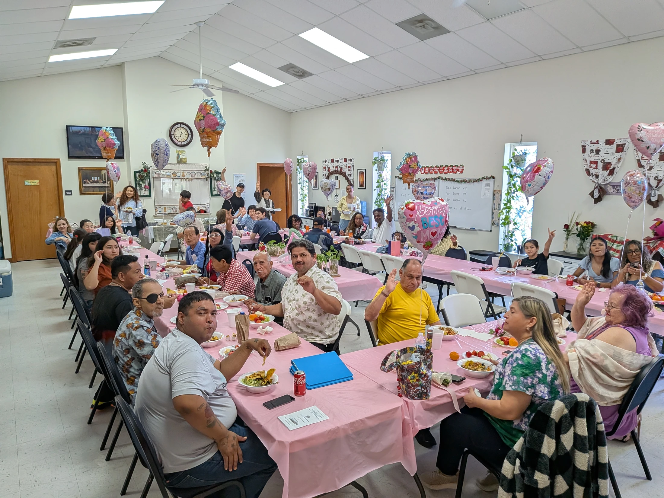 A large, diverse group of people celebrating Mother's Day at long tables with pink tablecloths in a decorated hall. Many are smiling and waving at the camera.