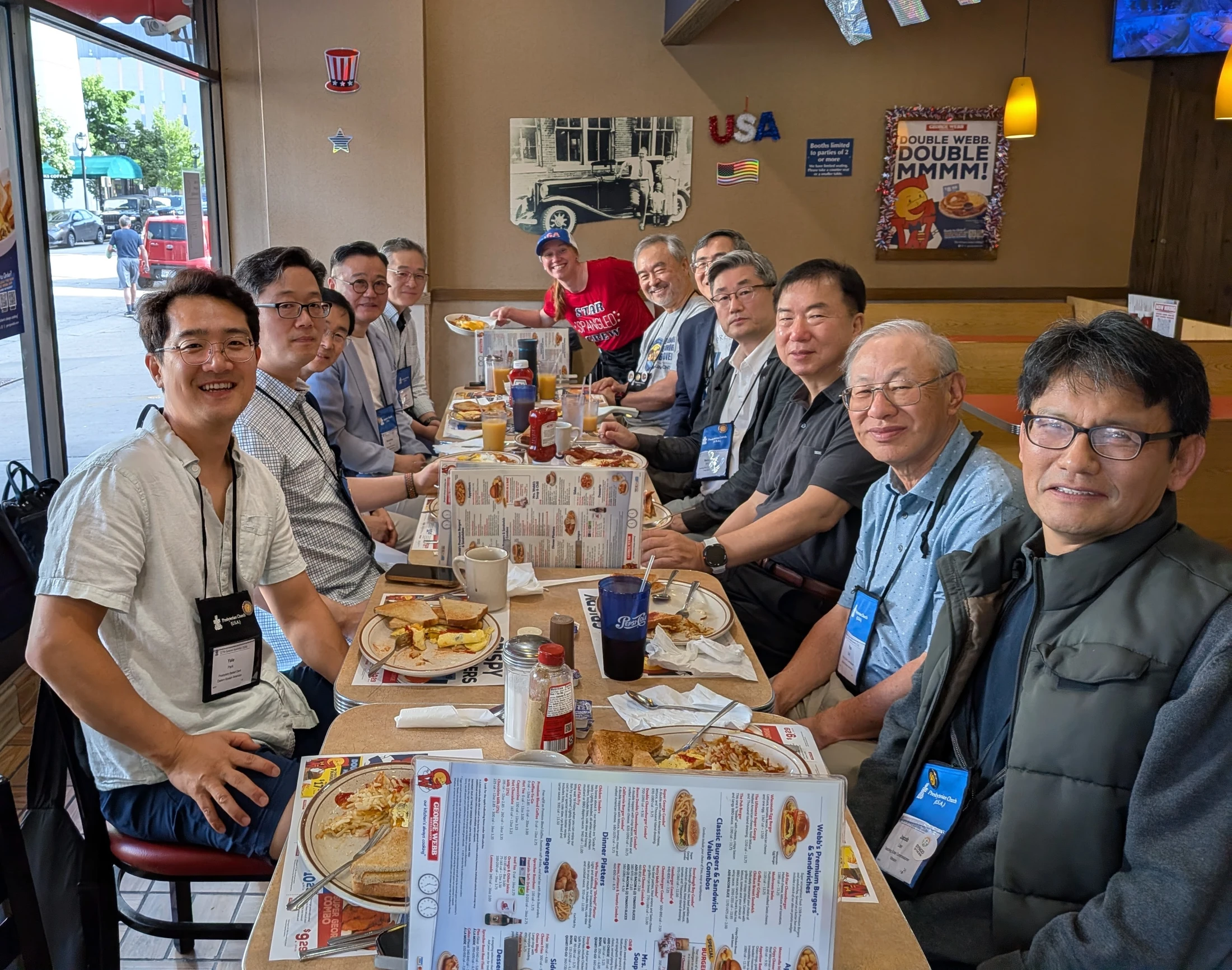 A large group of men wearing conference lanyards sit together at a long table in a diner, smiling and eating breakfast.