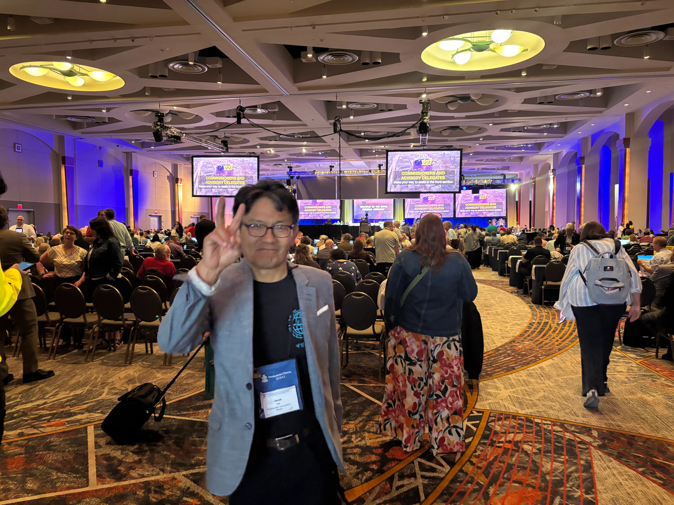 A man in a grey blazer and glasses smiles and makes a peace sign at the camera while standing in a crowded conference hall during a large assembly.
